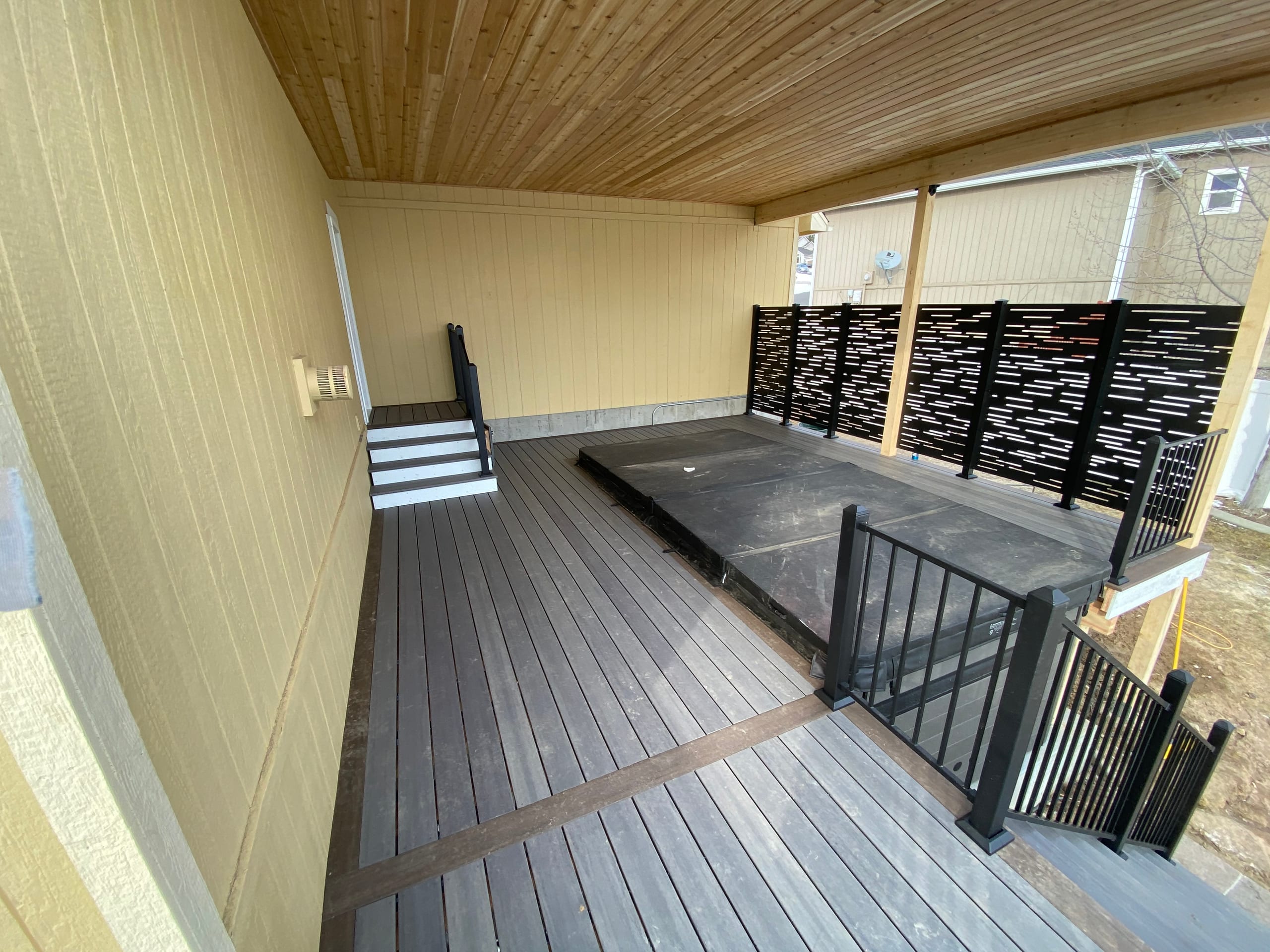 Inside a covered deck with cedar ceiling, dark grey composite decking, perforated privacy screen and black railing