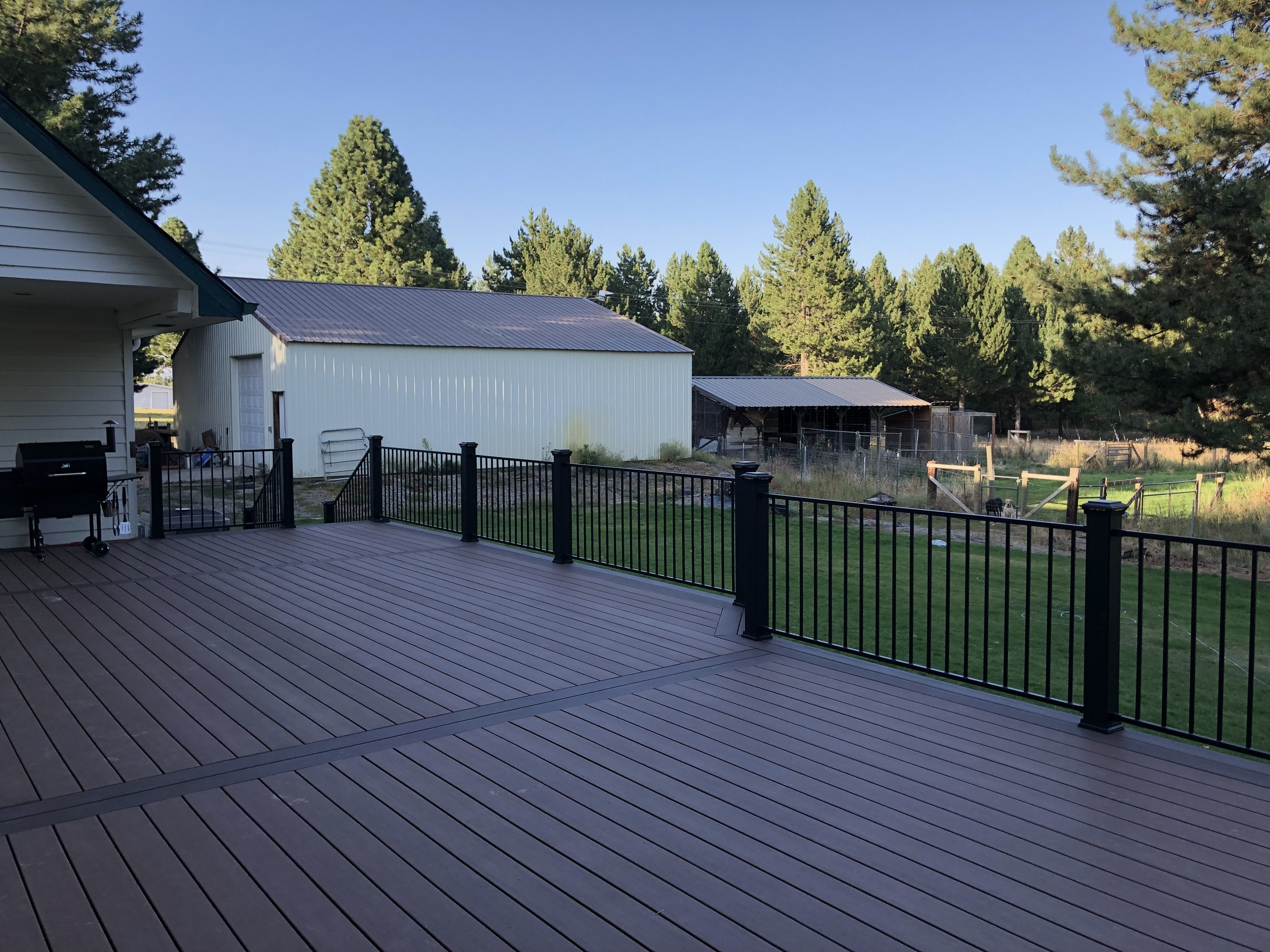 Large brown composite deck with inlay border, black railing, grill and a shop building and pines behind