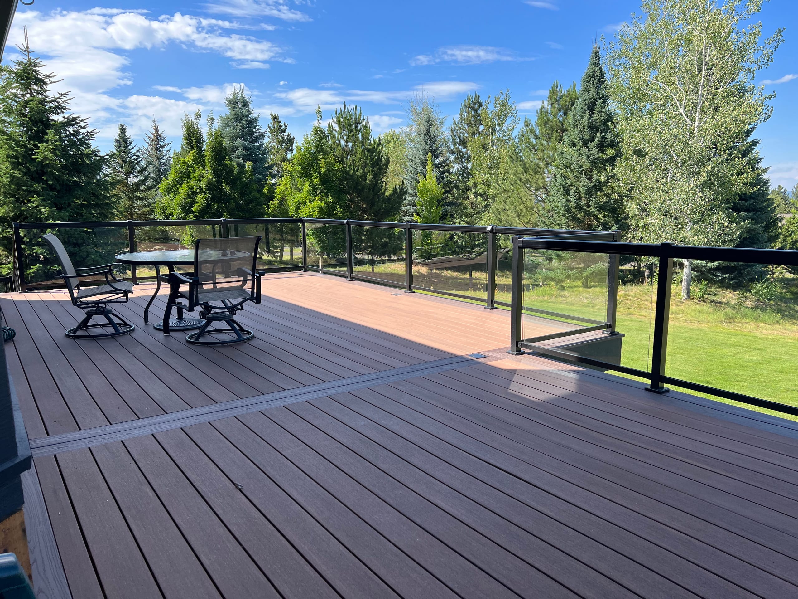 Large brown composite deck with patio sets, black frame glass-panel railing and lawn and tree views