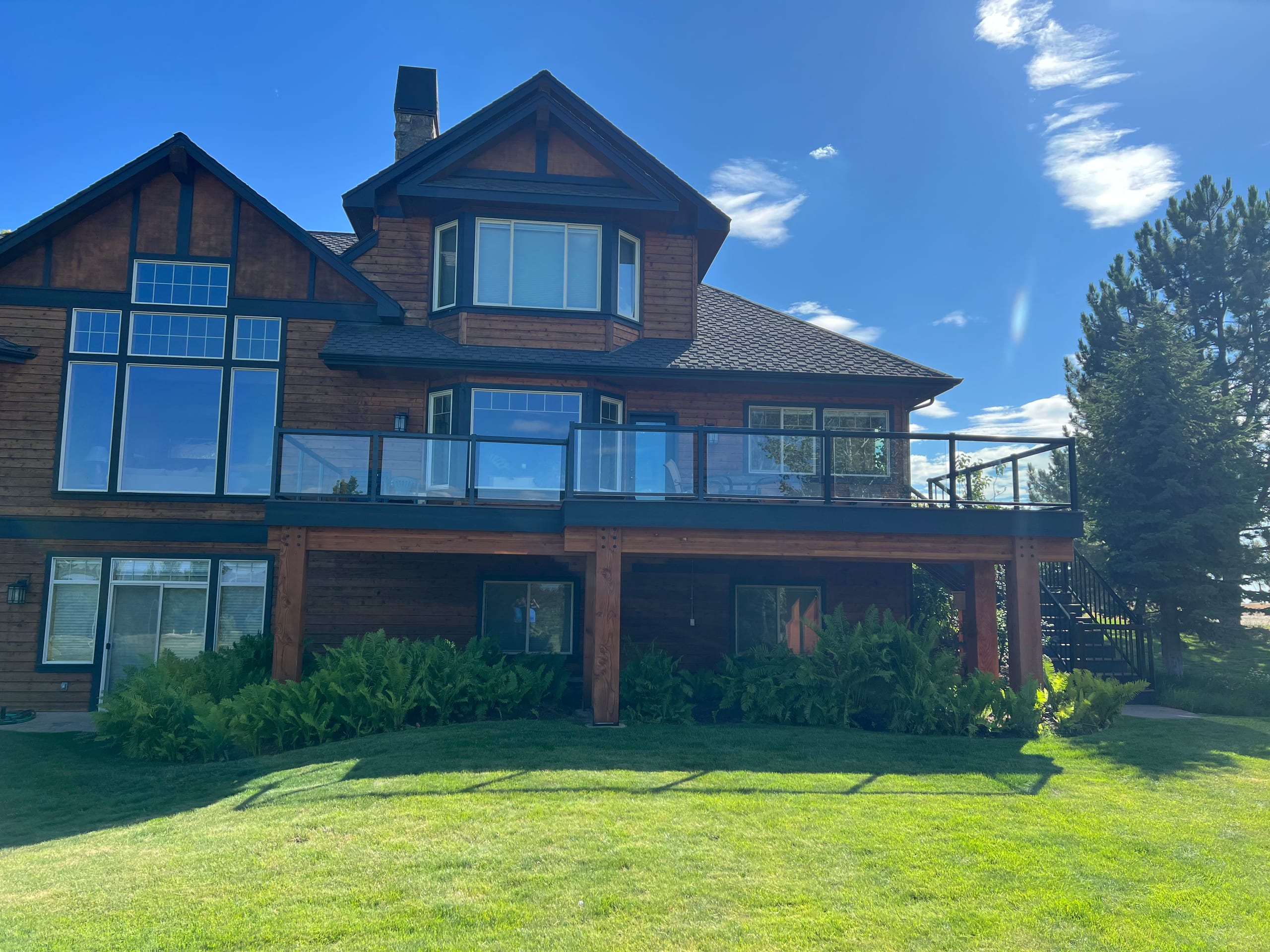 Large cedar-sided house with upper deck with glass railing, timber posts, big lawn and blue sky