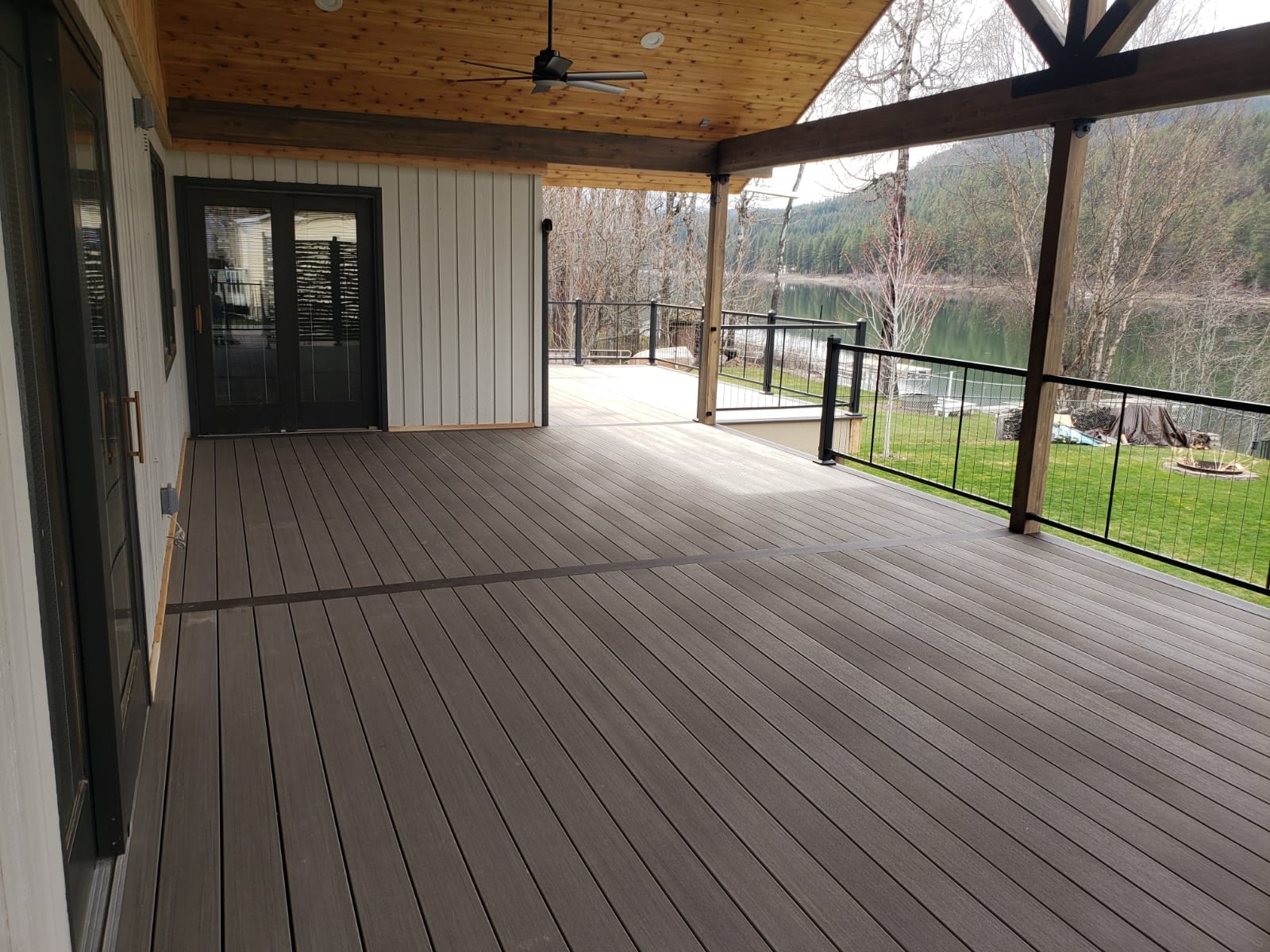 Large covered deck with brown composite decking, cedar ceiling with fan, black cable-style railing and lake view