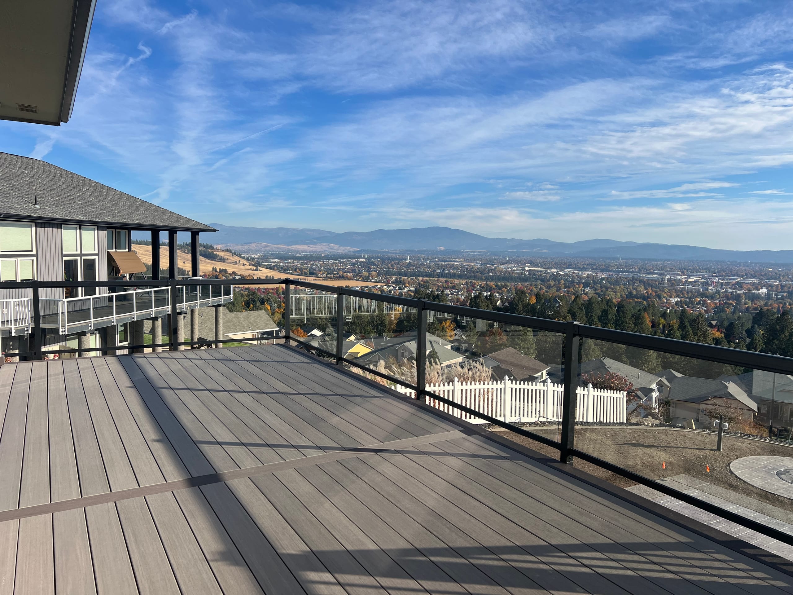 Large gray composite deck with glass railing and black frame overlooking Spokane valley and mountains under blue sky