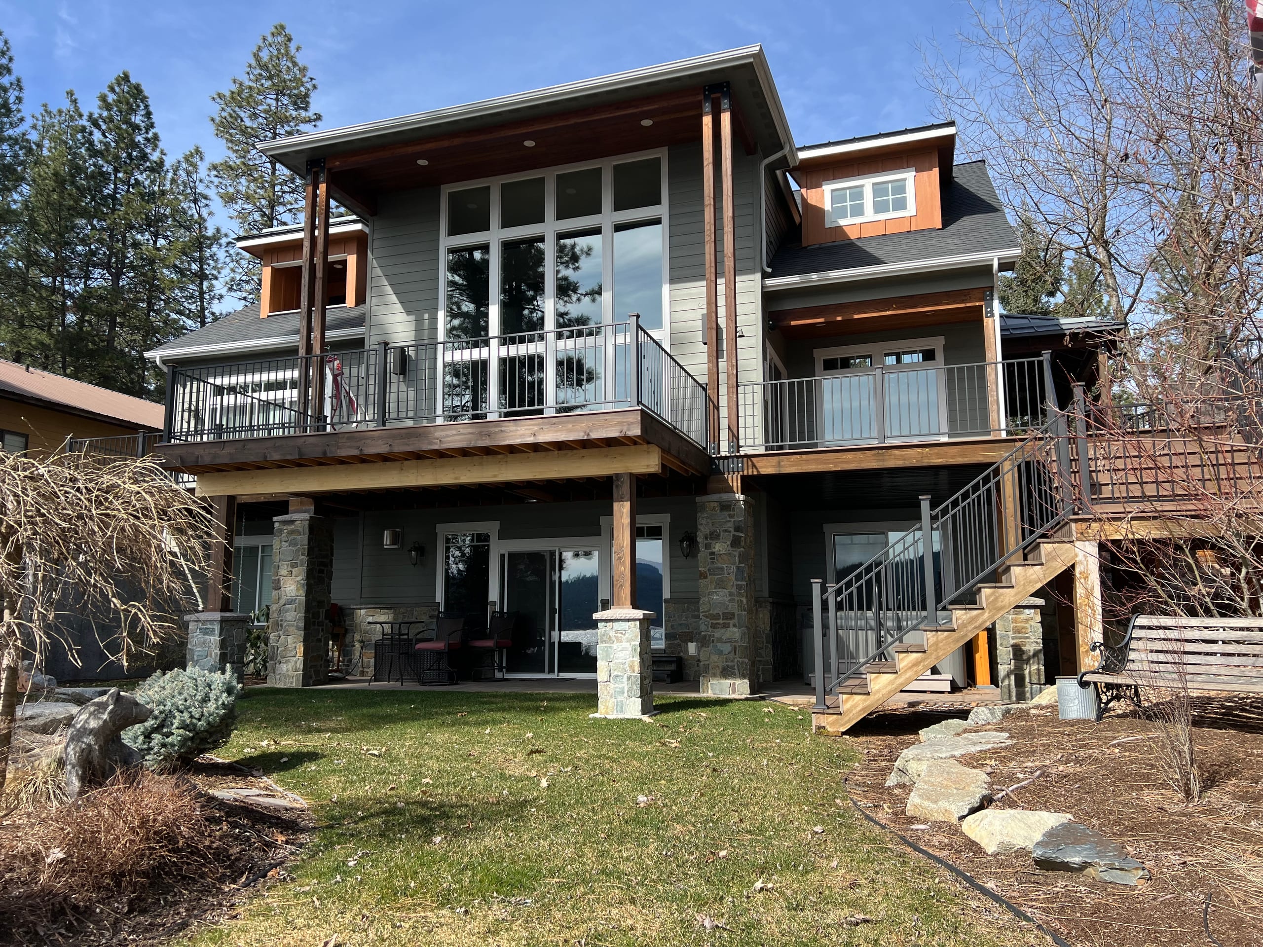 Large gray house with two-level decks with white railings, timber posts, stone columns and stairs over a lawn