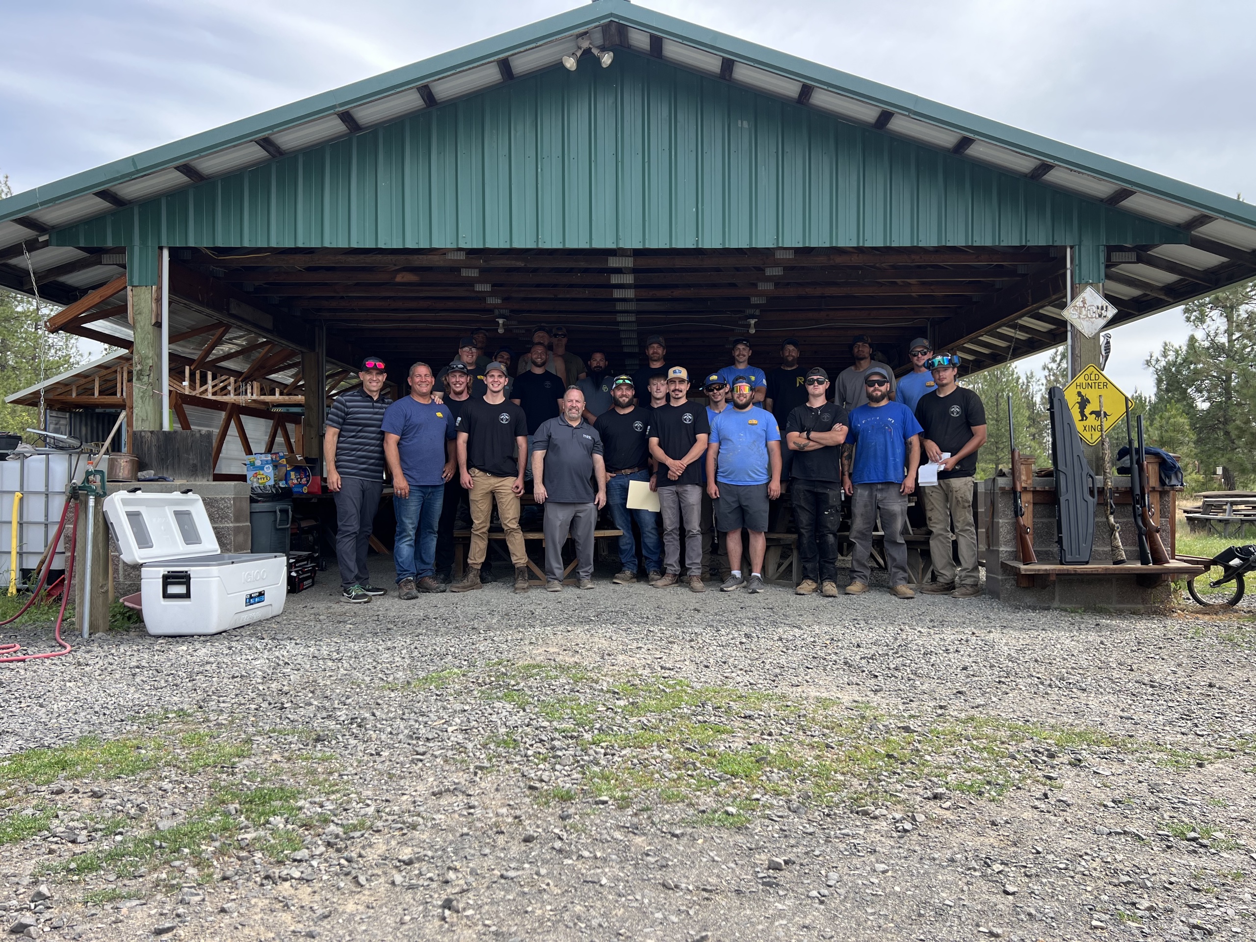 Large group photo of the crew standing in an open green-metal shed on a gravel lot