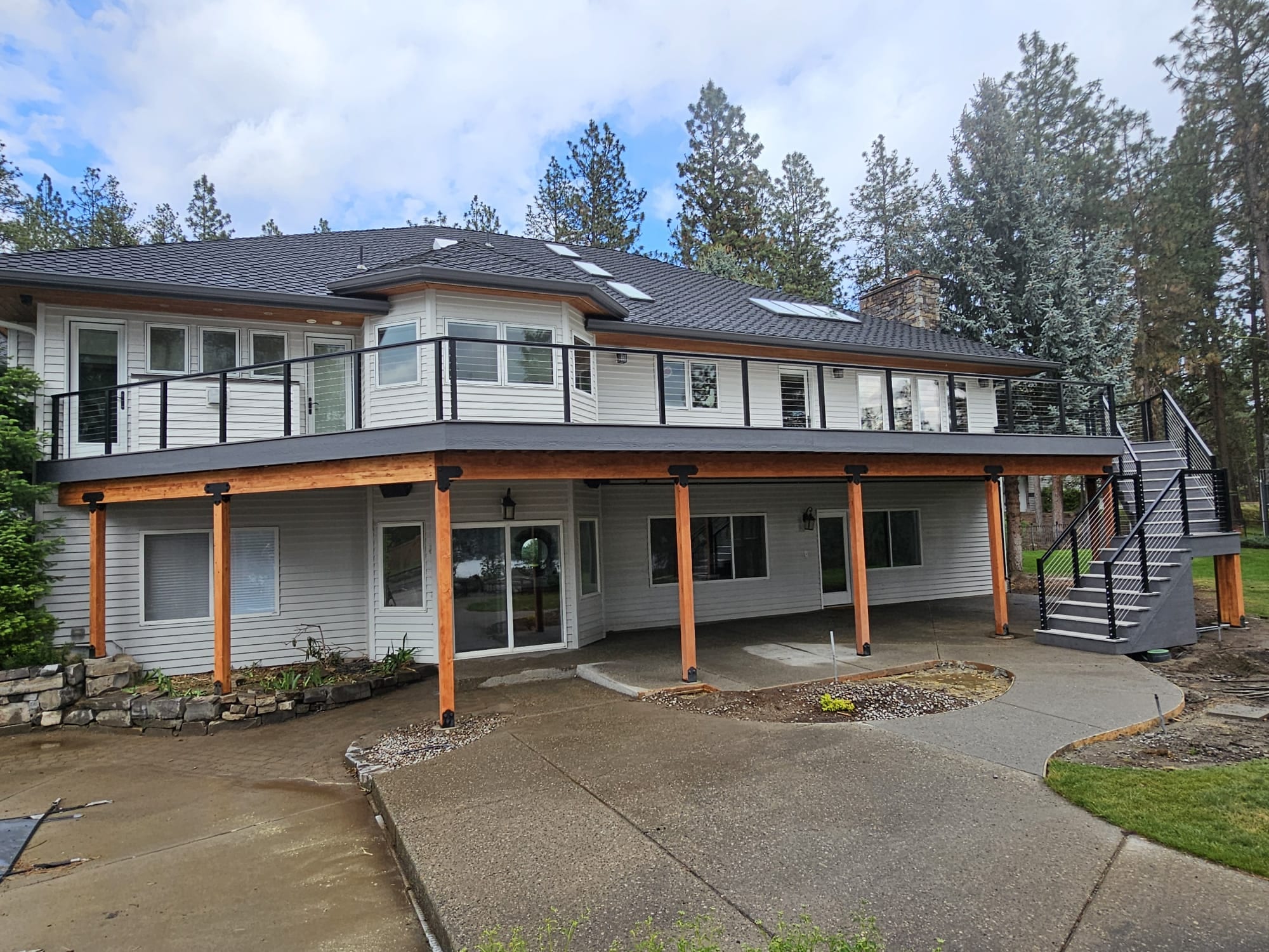 Large house with elevated deck on cedar posts, glass/cable-style black railing and covered lower patio, steps at right