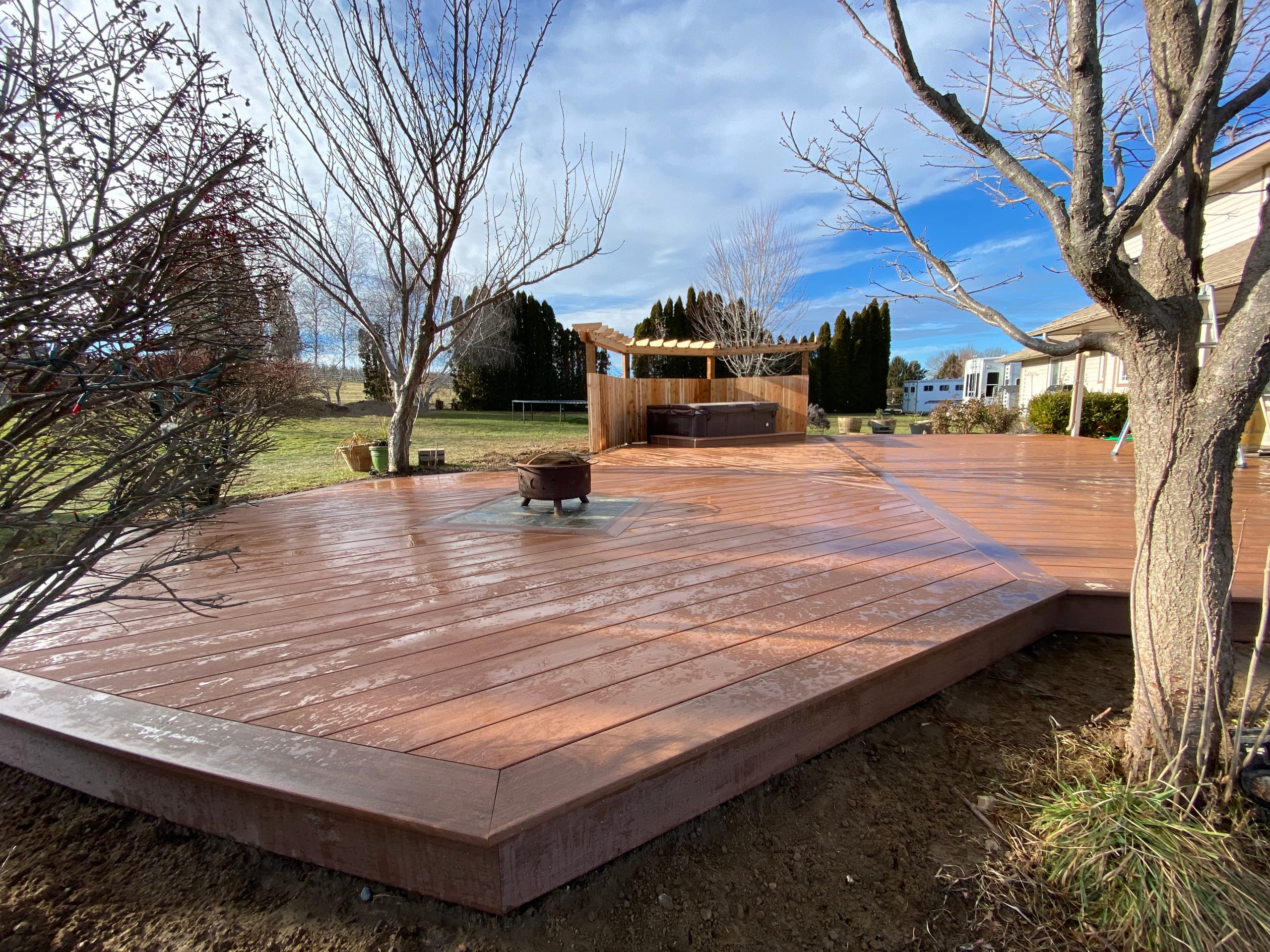 Large low-profile reddish composite deck with fire pit and cedar pergola at far end, bare trees and blue sky