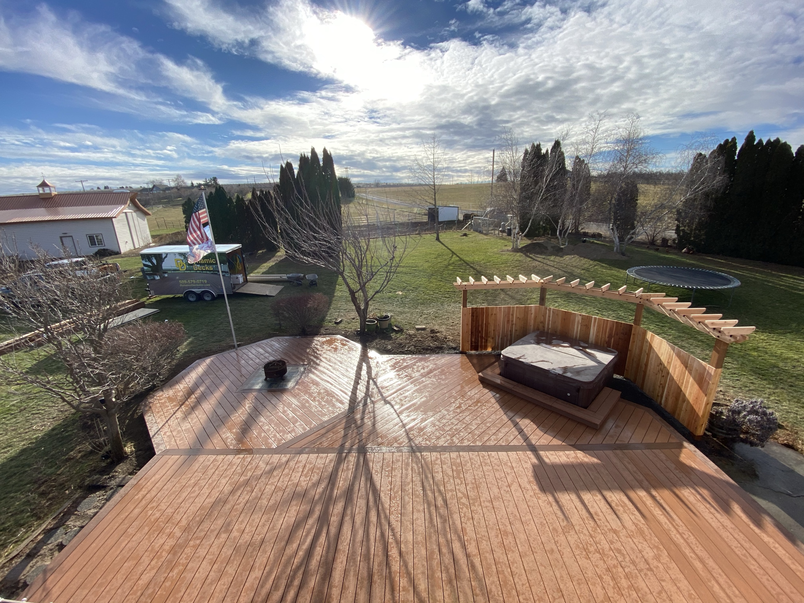 Large redwood-tone composite deck with built-in cedar pergola and hot tub, lawn and farm buildings, blue sky