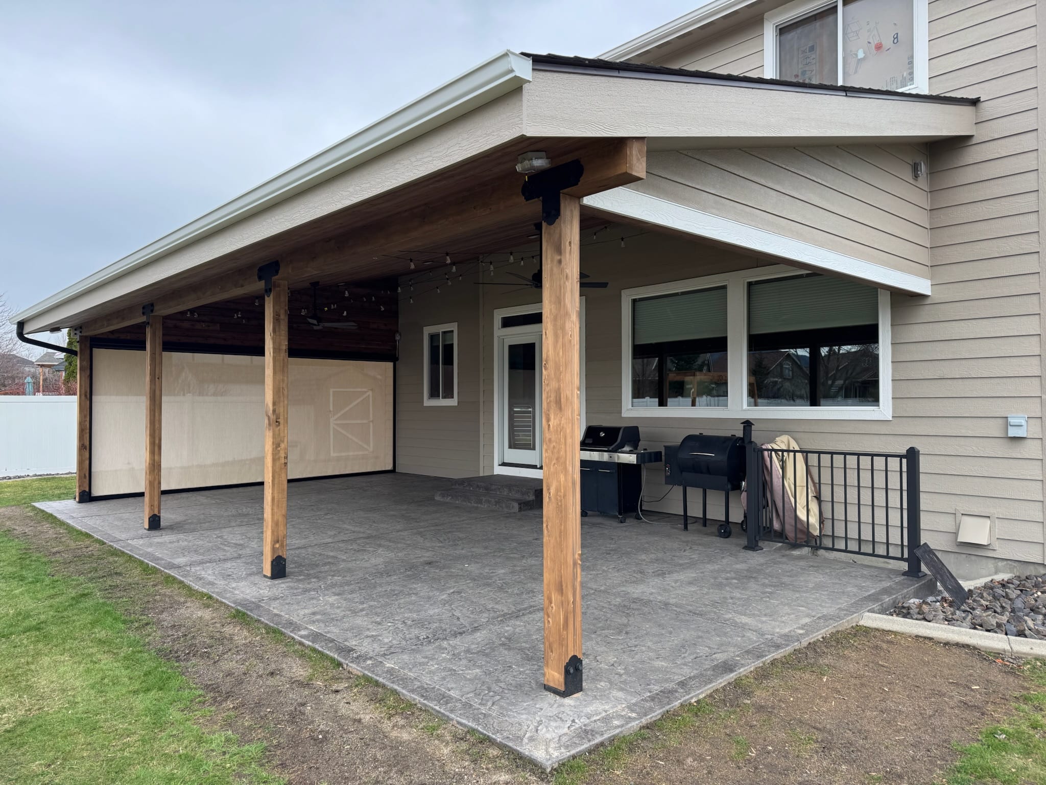 Large shed-roof patio cover on stained posts with black bases over stamped concrete, tan siding, grill and sun shades