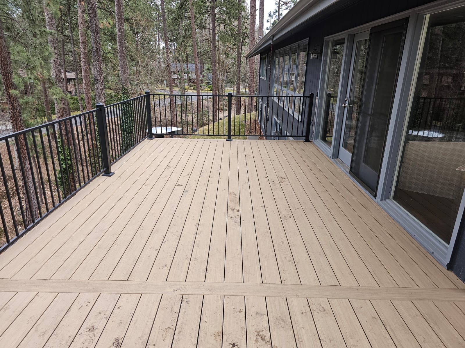 Large tan composite deck with black metal picket railing and sliding glass doors, pine forest behind