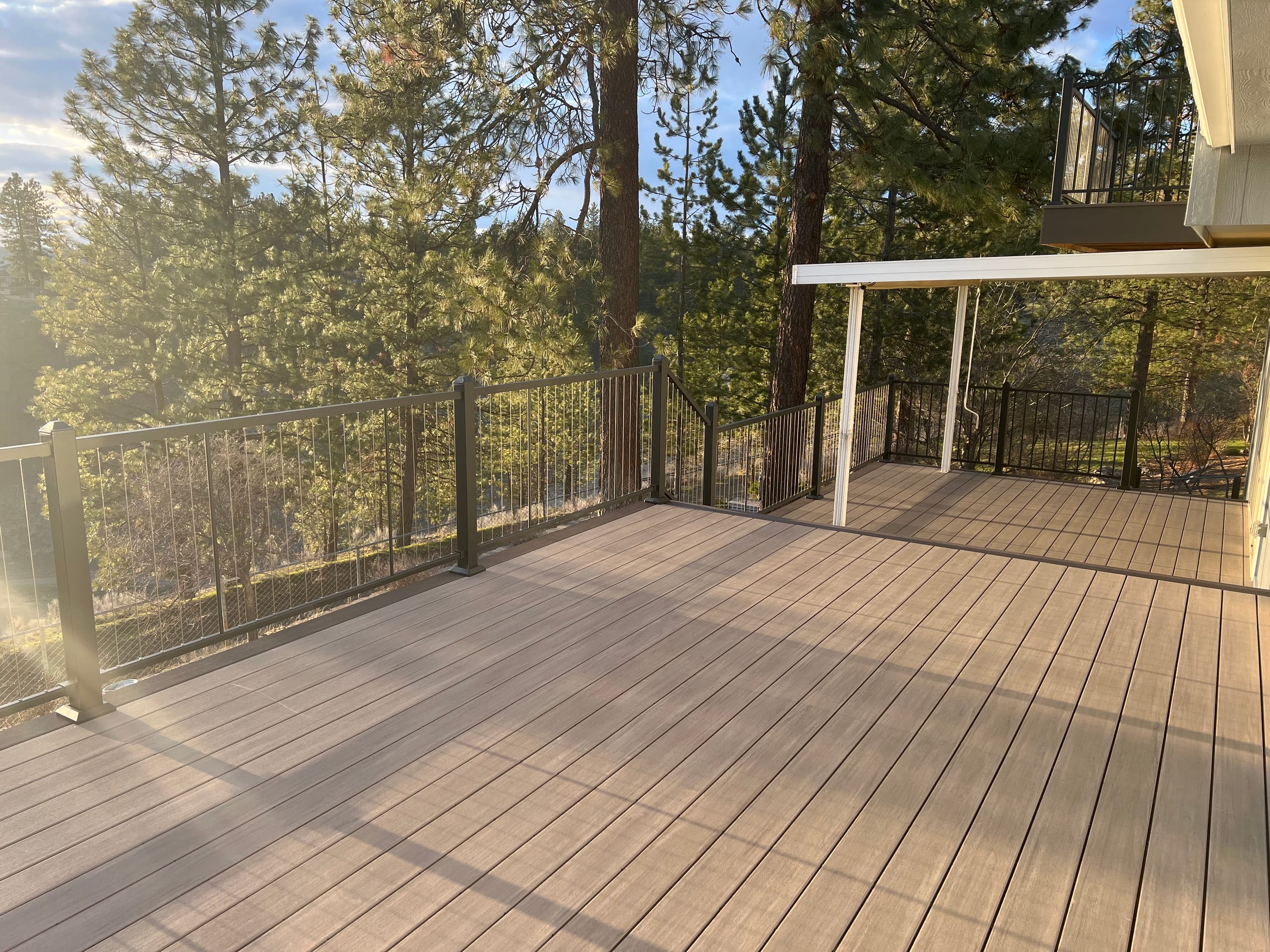 Large tan composite deck with black picket railing and aluminium patio cover in golden evening light over pine forest