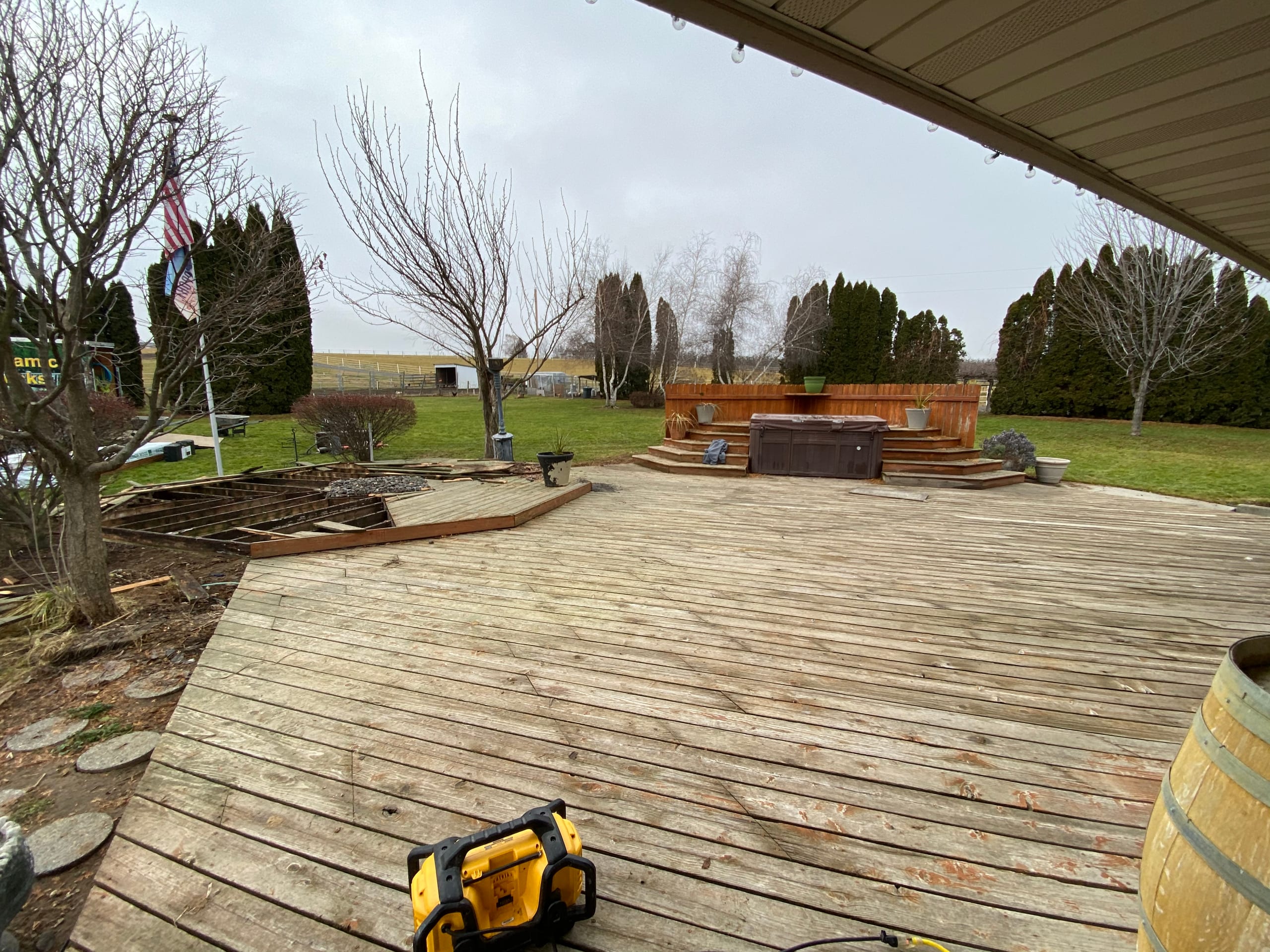 Large weathered grey wood deck with curved edge, hot tub beyond, DeWalt radio on boards, rural yard