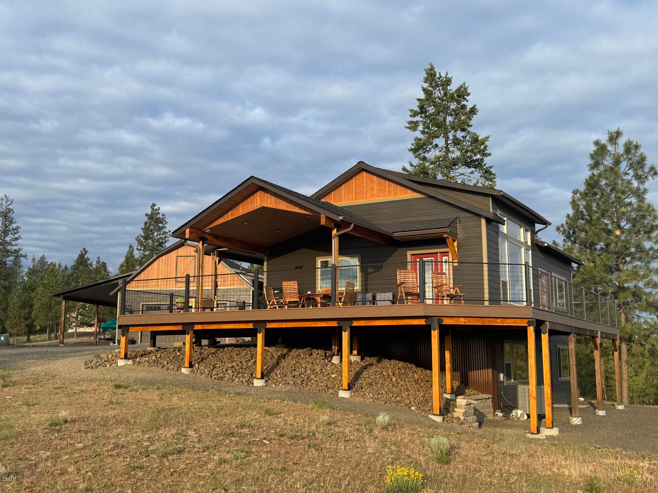 Large wraparound elevated composite deck with covered section on a dark gabled house, timber posts on a hillside with pines