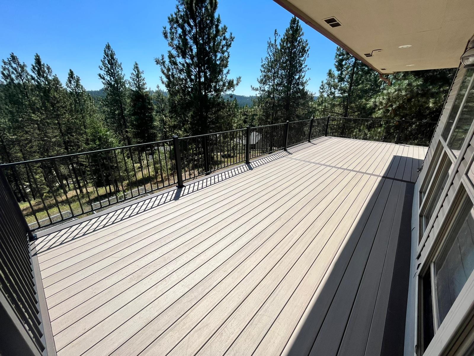 Light gray composite deck with black railing and tall pine forest views, under a house eave