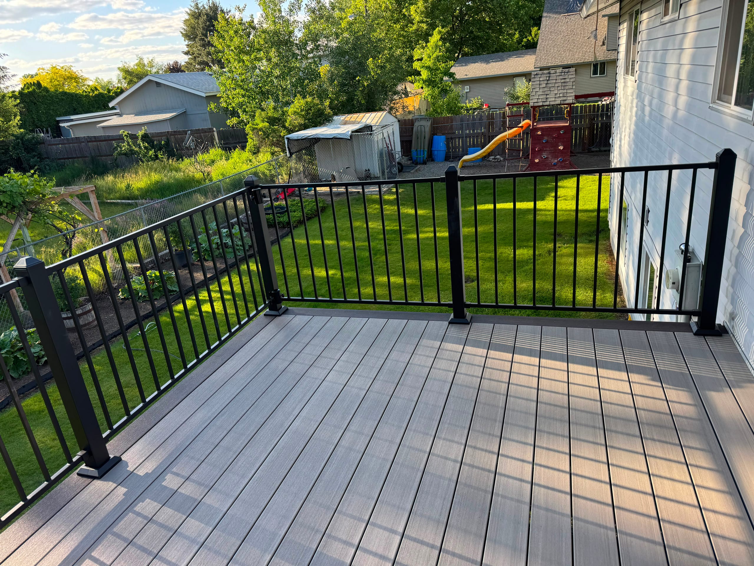 Light grey composite deck with black metal picket railing beside white house, green lawn and play set beyond