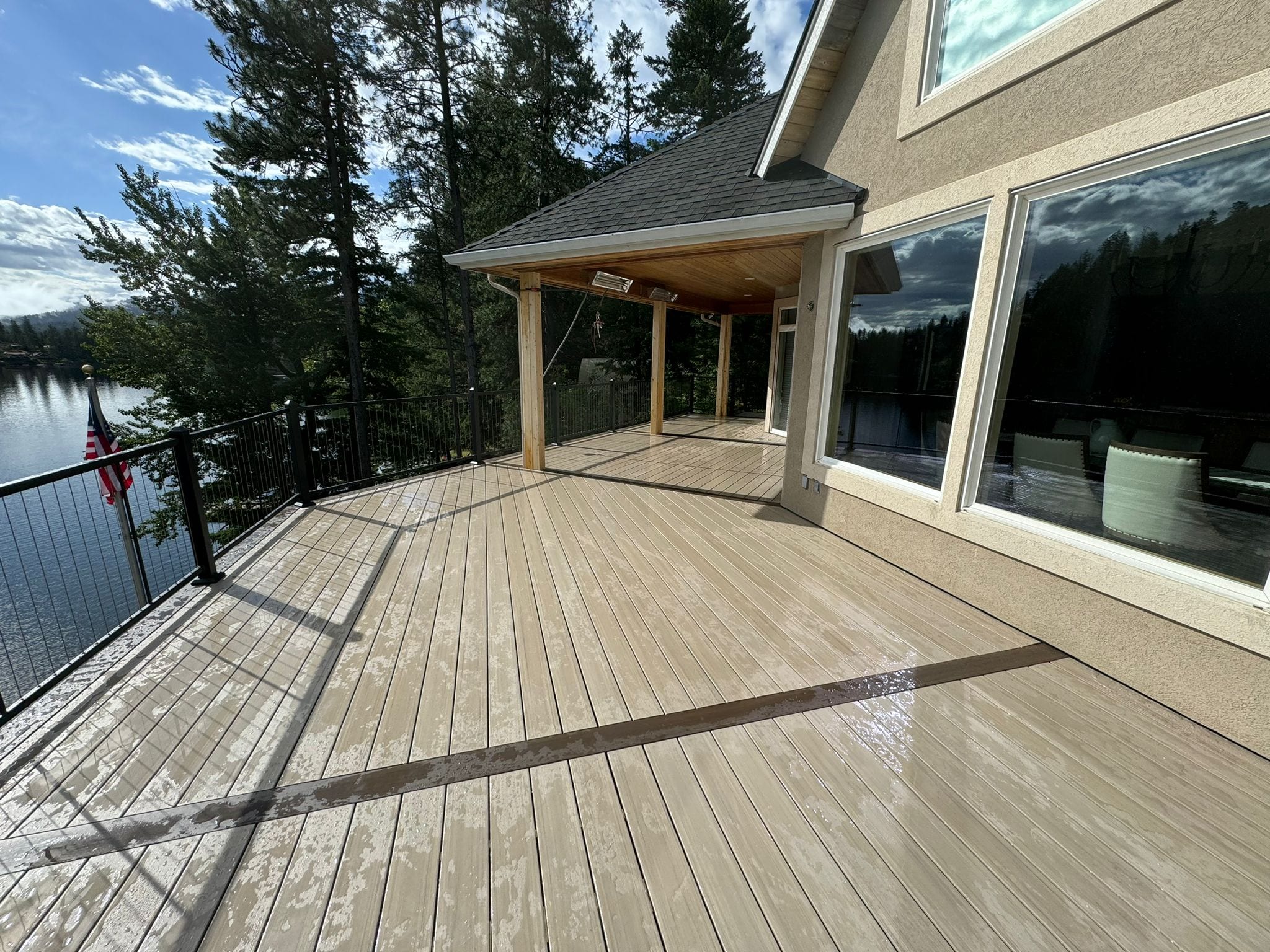 Light tan composite deck with dark inlay band, partial wood-ceiling roof, large windows and black railing over a lake