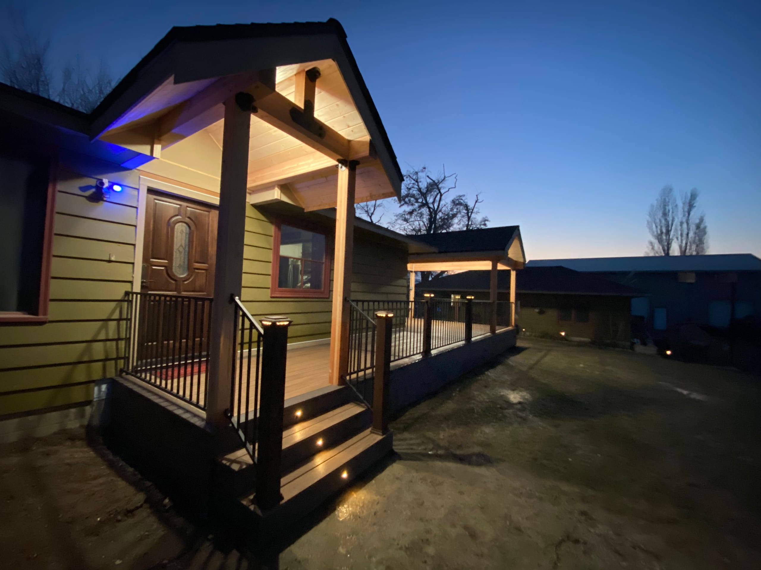 Lit gabled entry porch with timber posts, cedar ceiling, ornate door, black railing and lit steps at blue hour