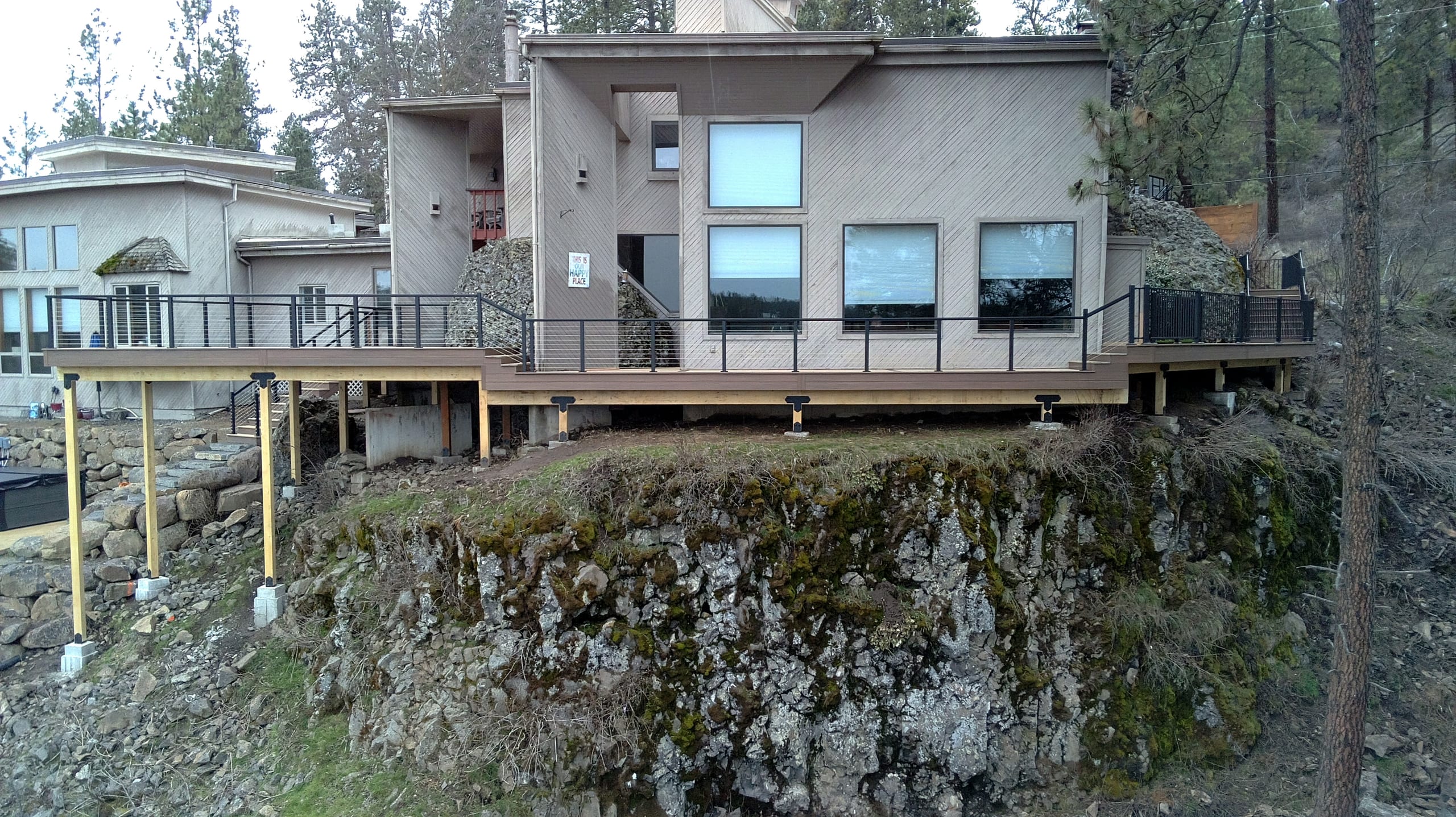 Long elevated deck on tall posts over a rocky bluff behind a tan stucco house, black railing, wood framing