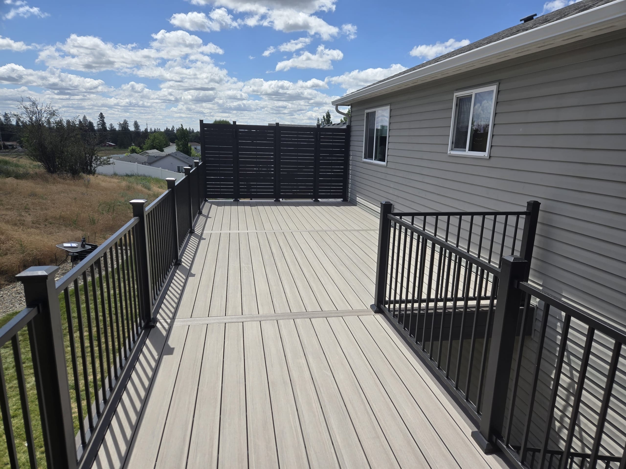Long light gray composite deck with black metal picket railing and black slat privacy screen at the far end, gray house