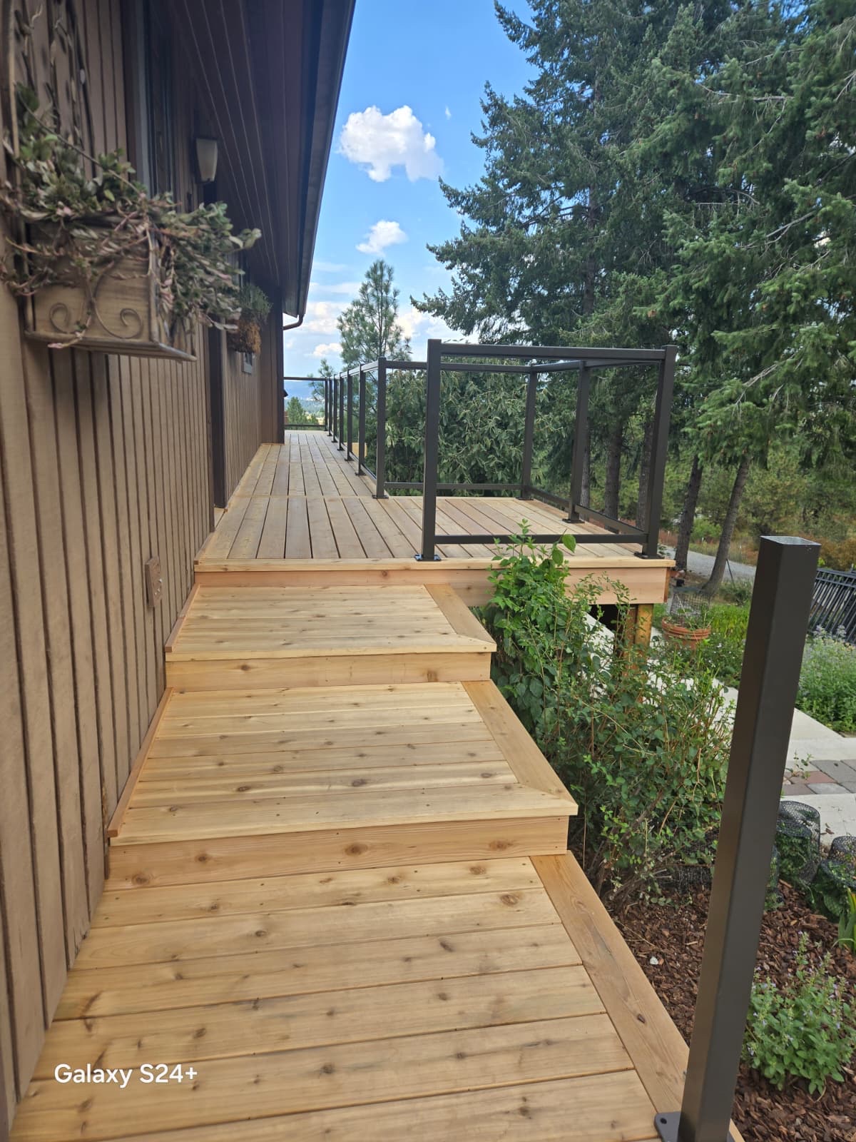 Long narrow cedar wood walkway deck with steps along house side, black railing at end, trees beyond