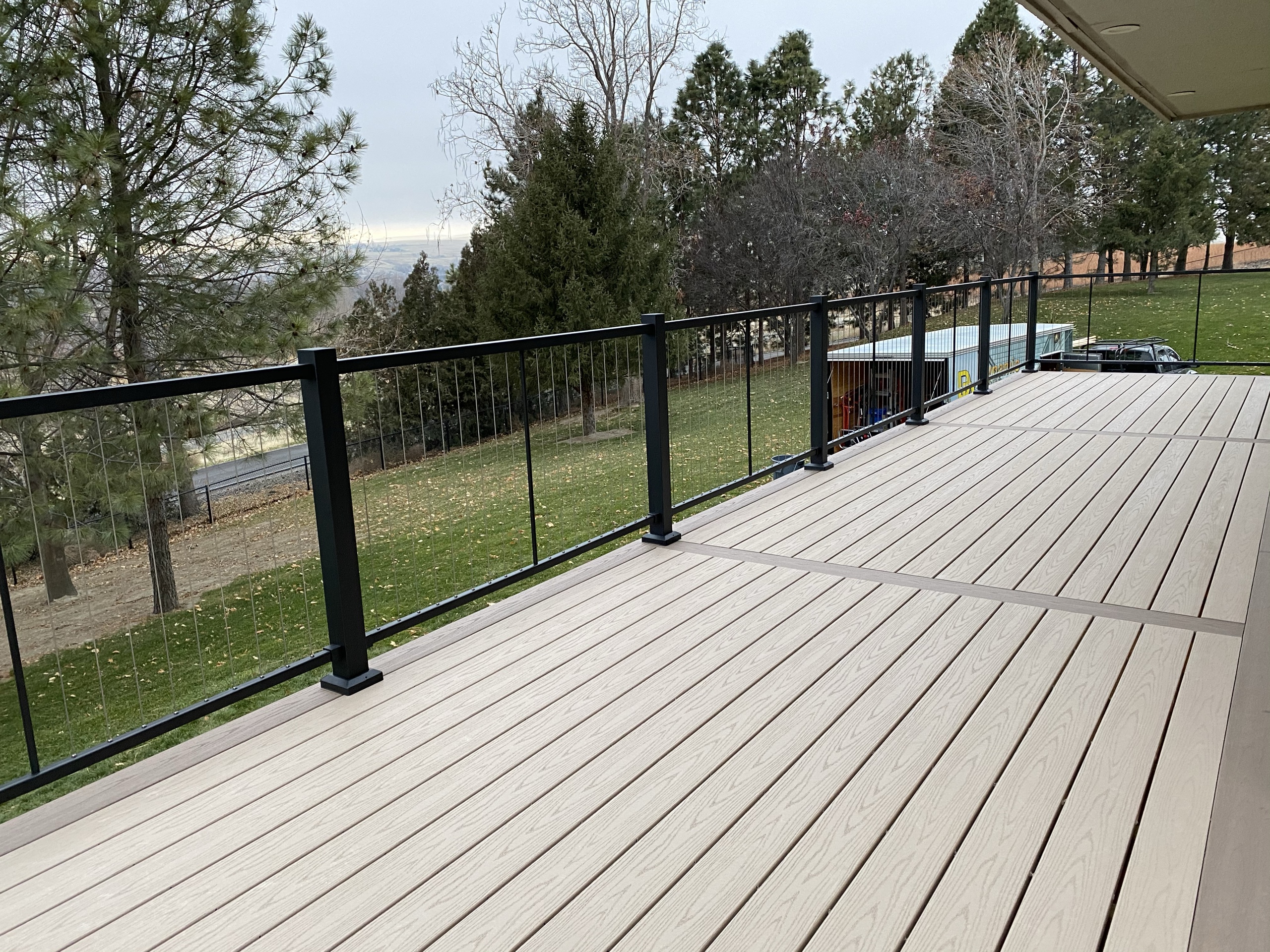Long tan composite deck with black cable railing overlooking green field and trees, overcast