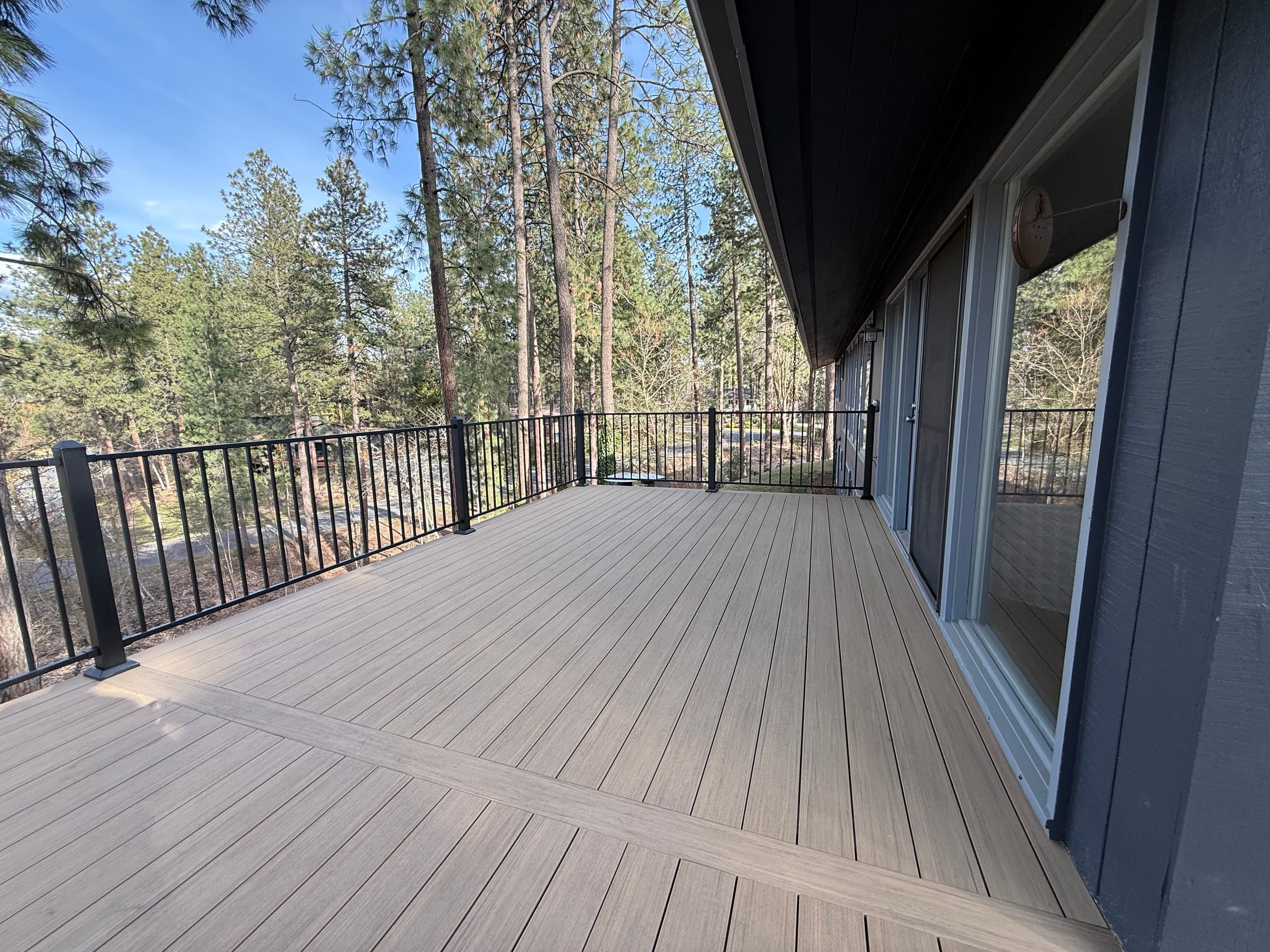 Long tan composite deck along dark house wall with black metal picket railing, pine trees beyond