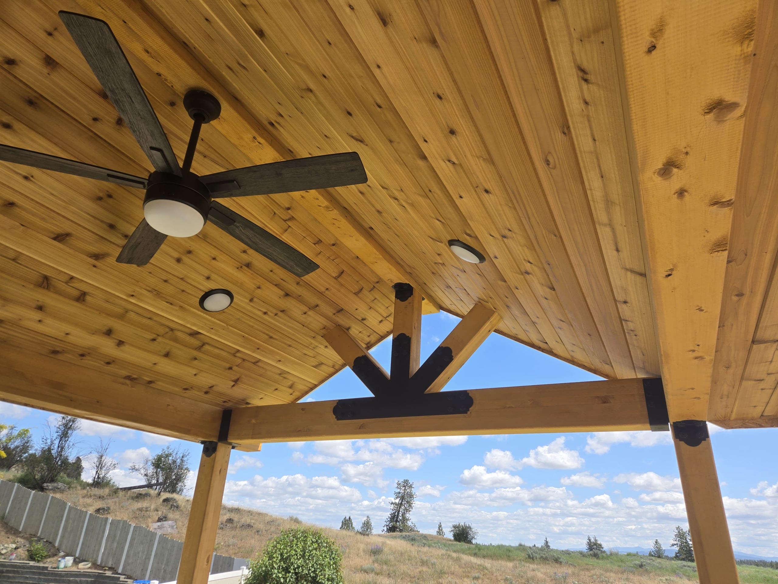 Looking up at gabled cedar plank ceiling with ceiling fan, recessed lights and dark steel truss bracket, valley view