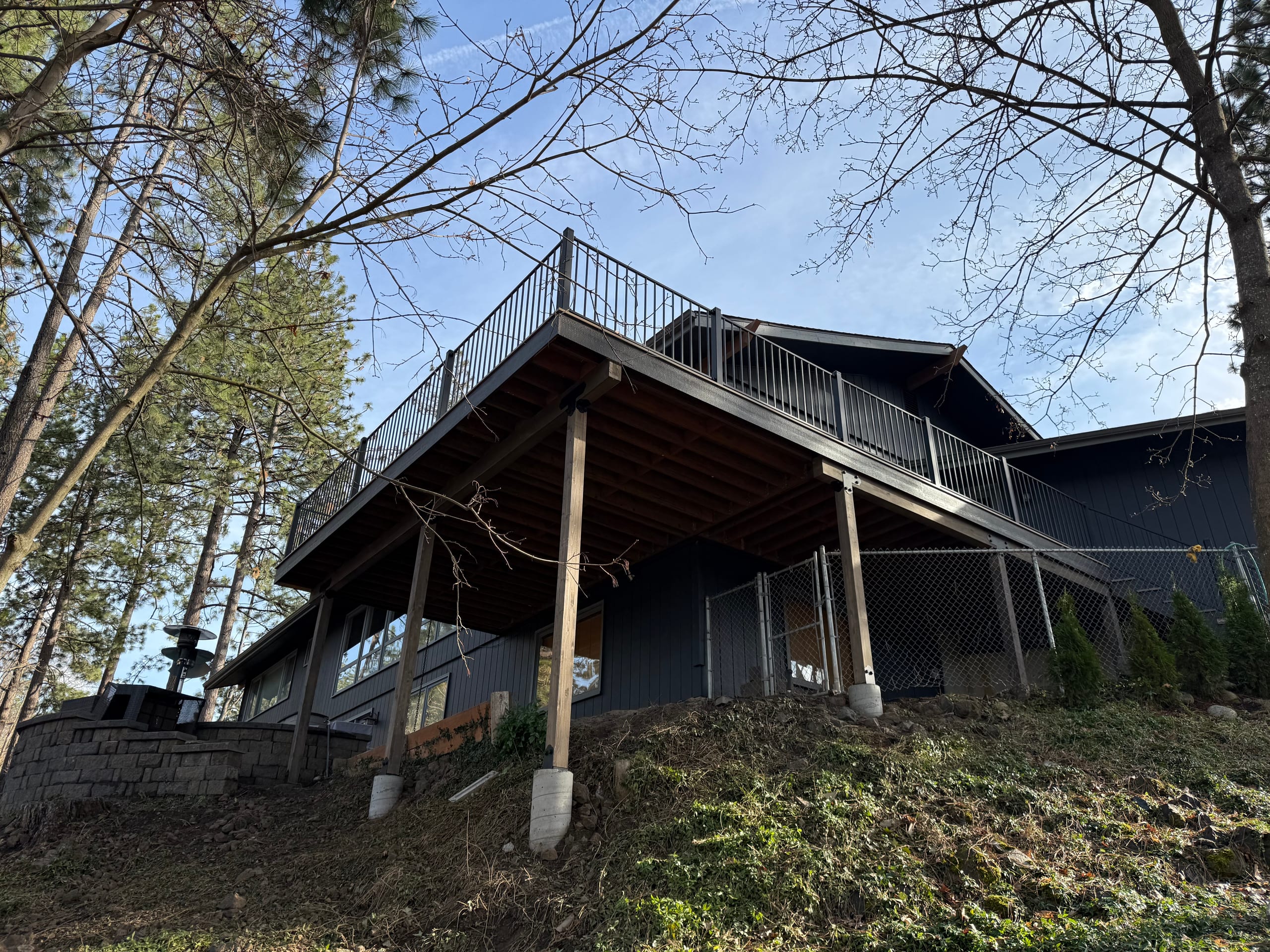 Low-angle view of large elevated deck with metal railing on tall posts and concrete footings up a hillside, dark house