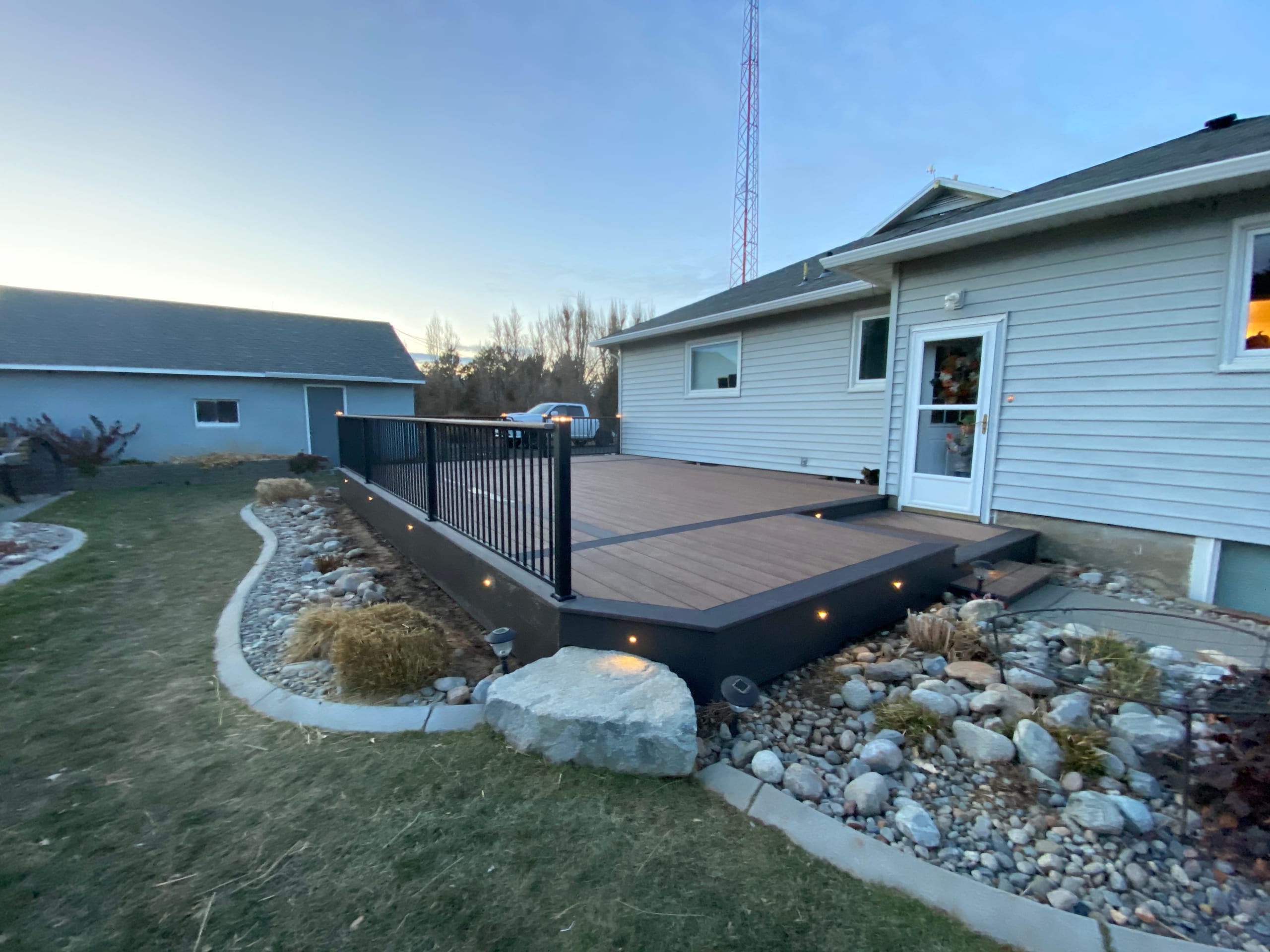 Low brown composite deck with black railing and recessed step lights at dusk beside blue house and rock landscaping