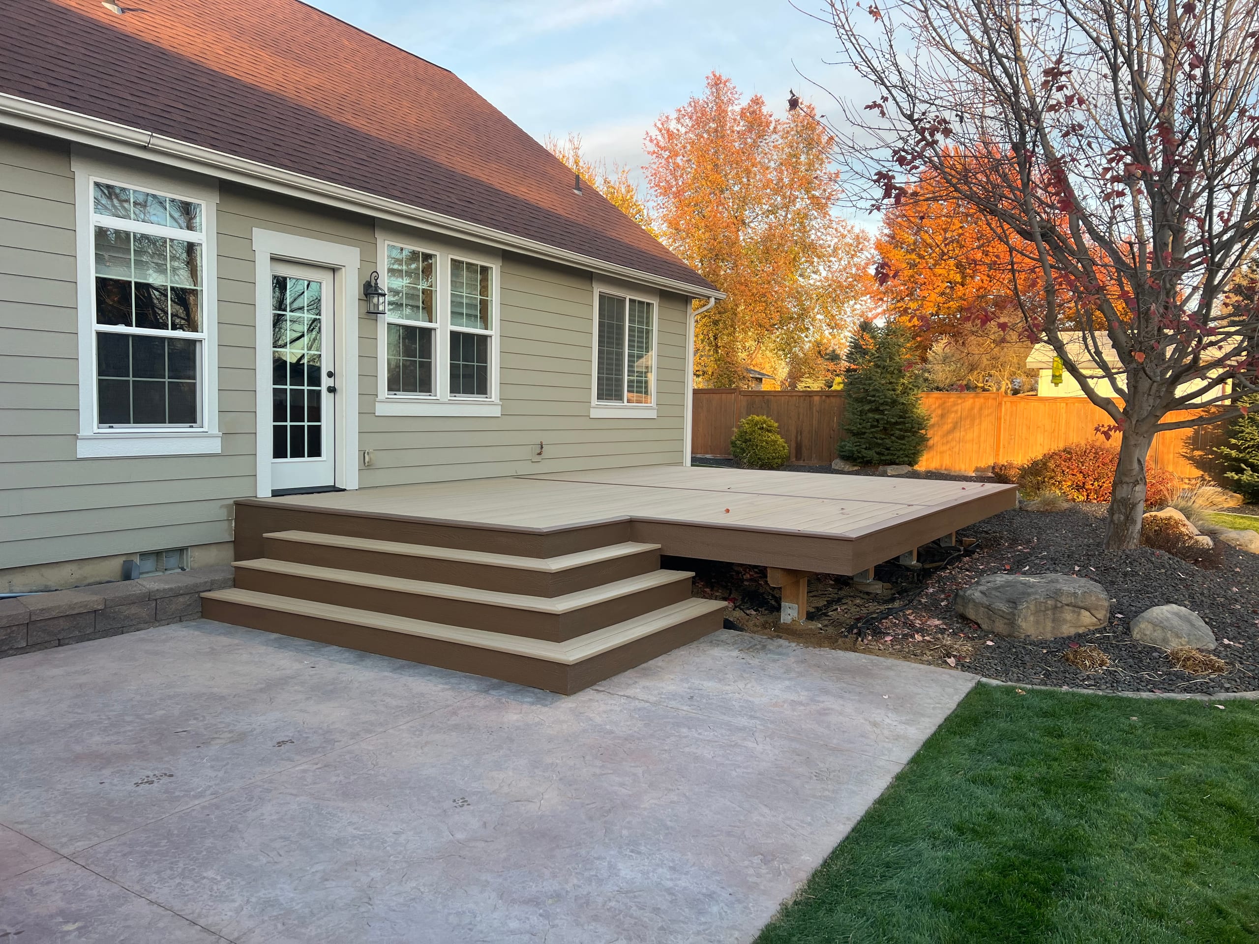 Low brown composite deck platform with wide steps down to a stamped concrete patio beside a tan house, autumn trees