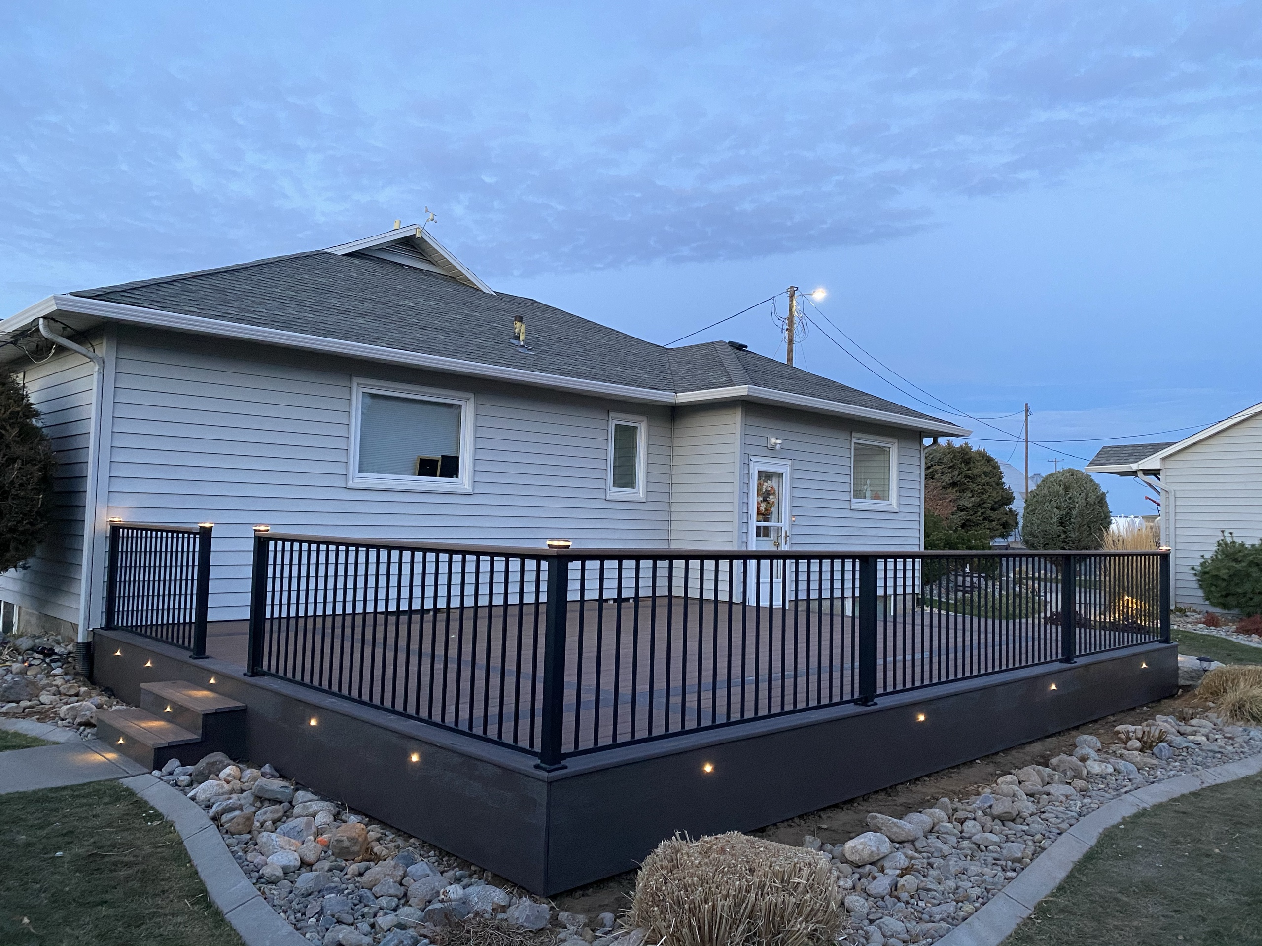 Low composite deck with black picket railing and lit fascia in twilight, curved rock border and lawn