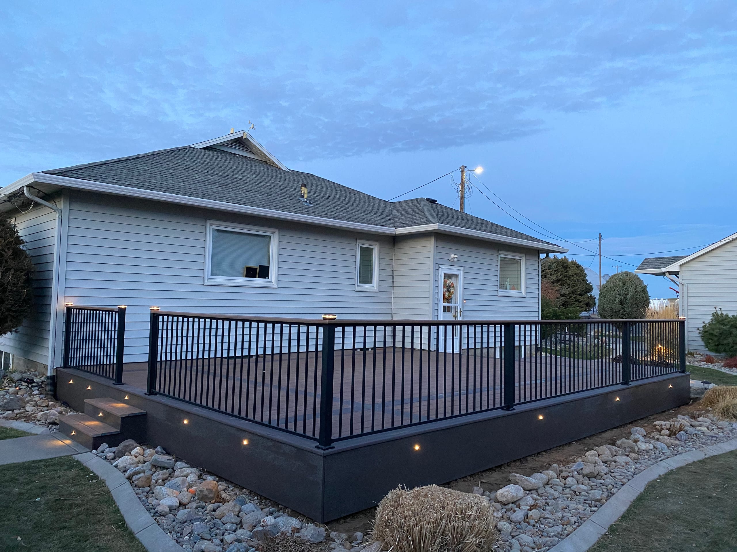 Low composite deck with black picket railing and lit fascia in twilight, curved rock border and lawn