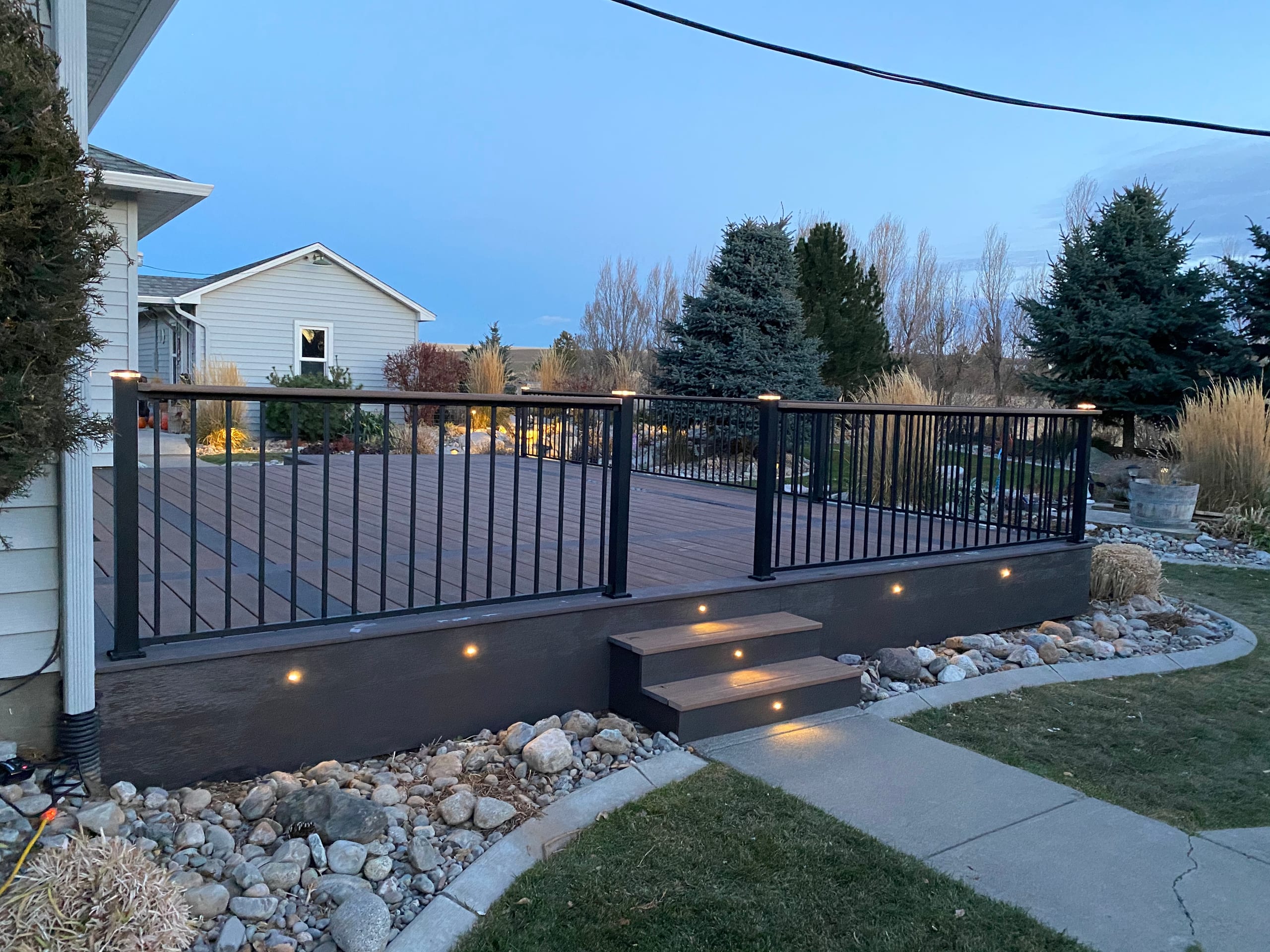 Low composite deck with black railing, lit steps and fascia lights at twilight beside pale house, rock-edged lawn