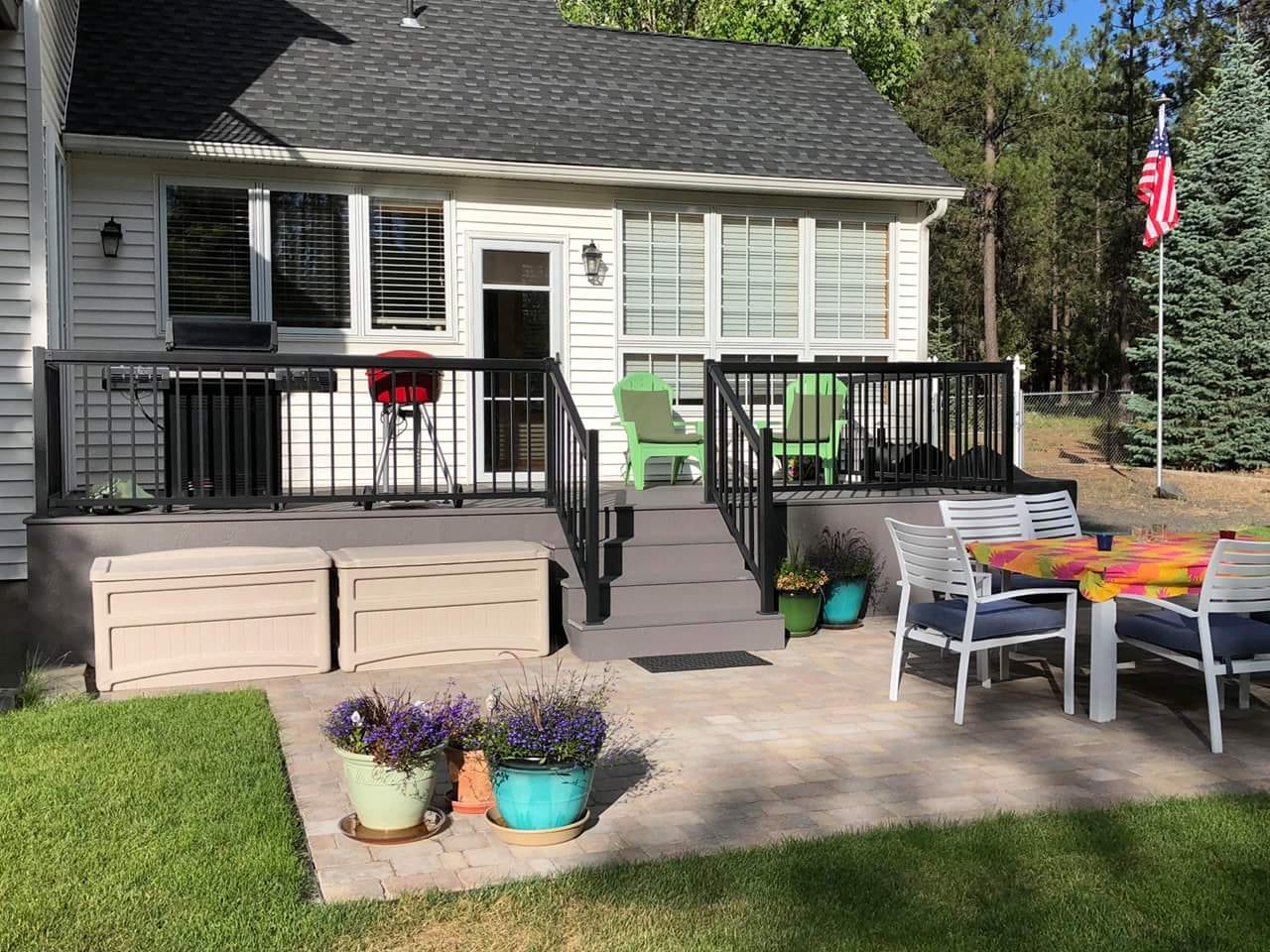 Low gray composite deck with black railing and stairs beside a paver patio with dining set and flower pots