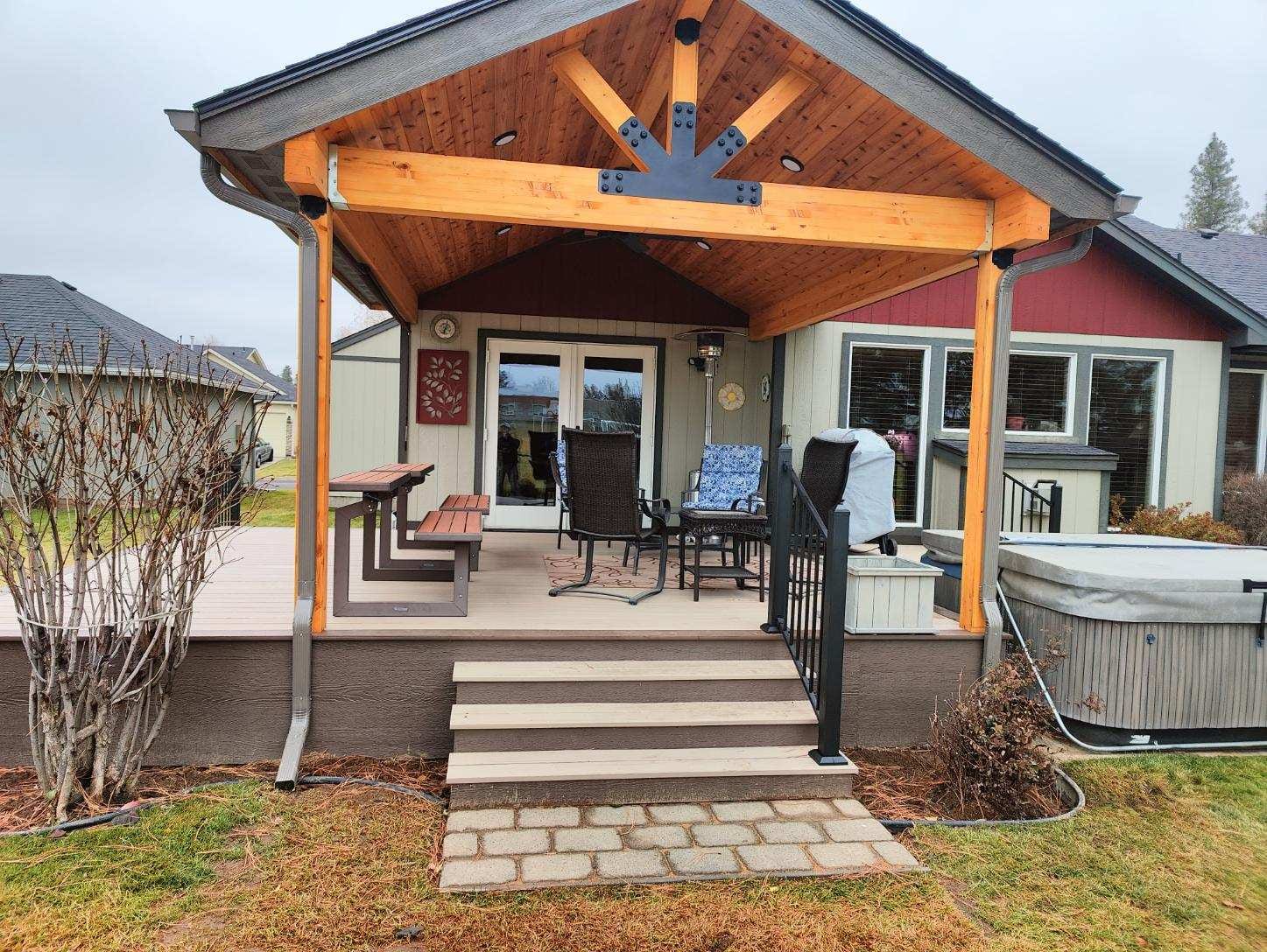 Low gray composite deck with gable cover, stained posts, black railing, hot tub and wide paver steps in front of tan house