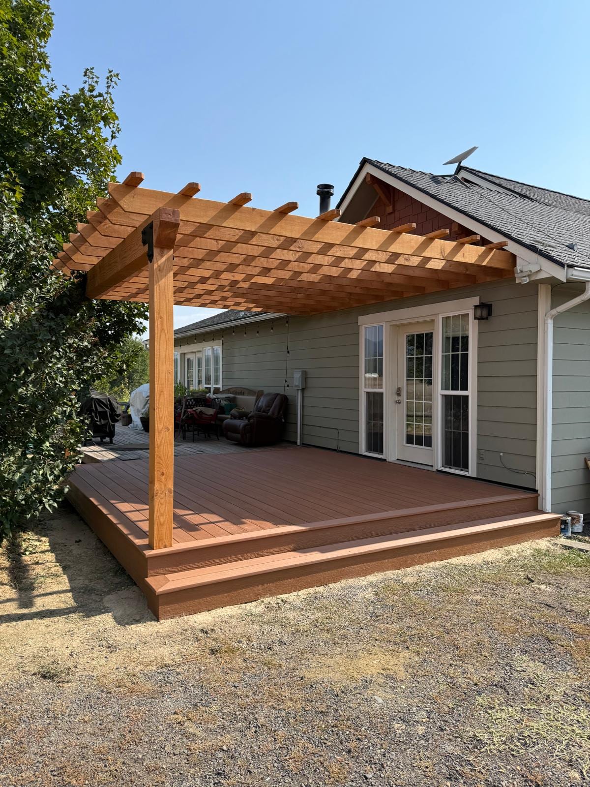 Low reddish-brown composite deck with cedar pergola attached to a gray house, dry lawn around