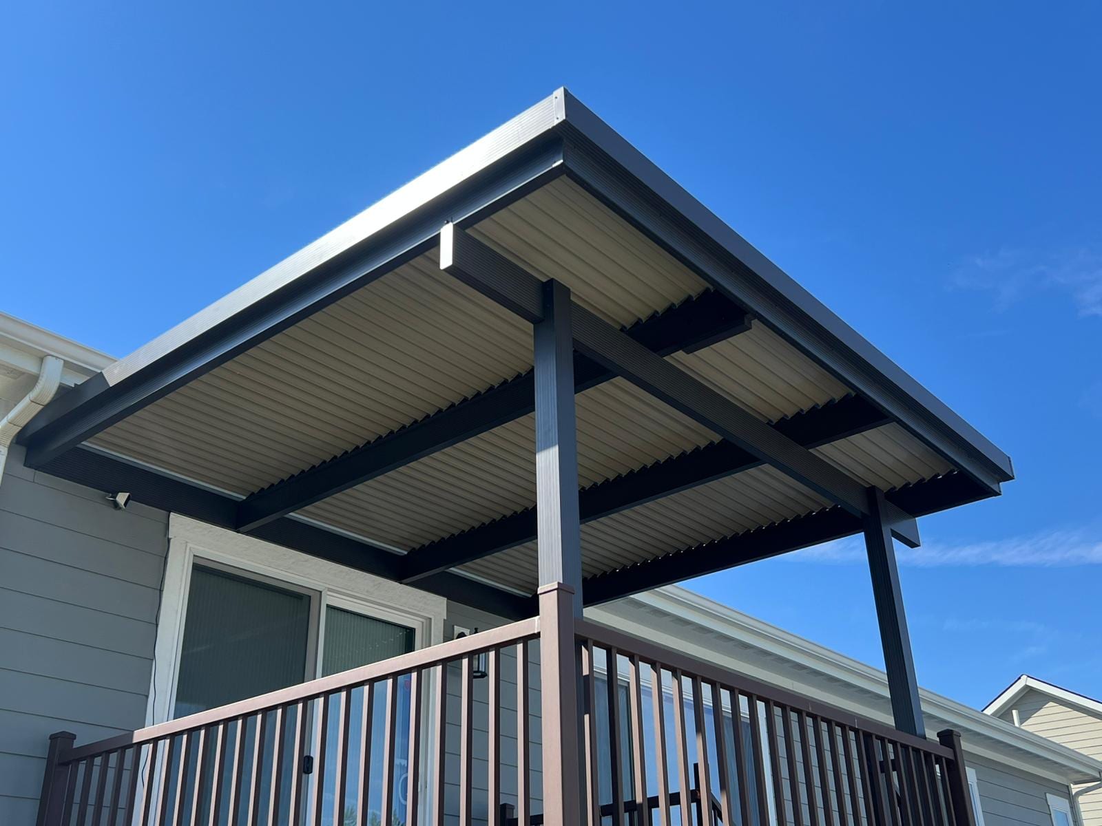 Modern flat metal-roof patio cover with dark steel posts over a deck with black picket railing, blue sky