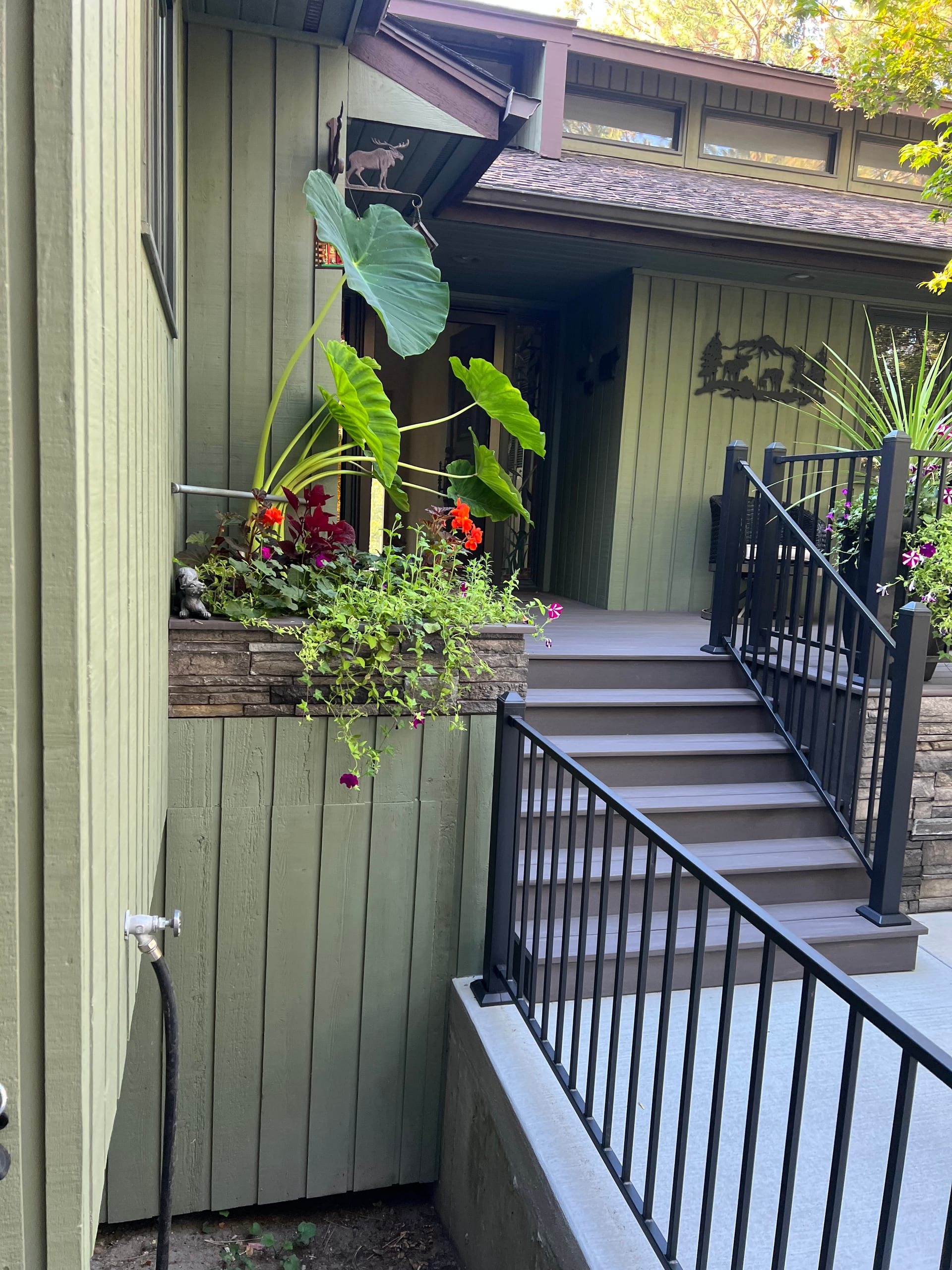 Narrow composite stair run with black railing beside green siding and stacked-stone planter with plants