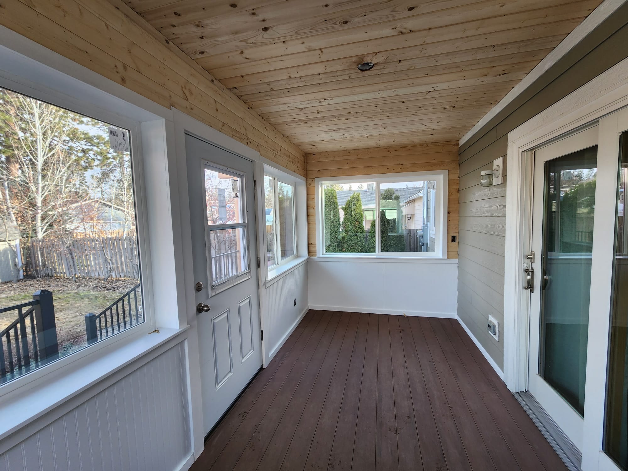Narrow enclosed room interior with wood ceiling, white wainscot, windows and dark composite floor, door and slider at sides