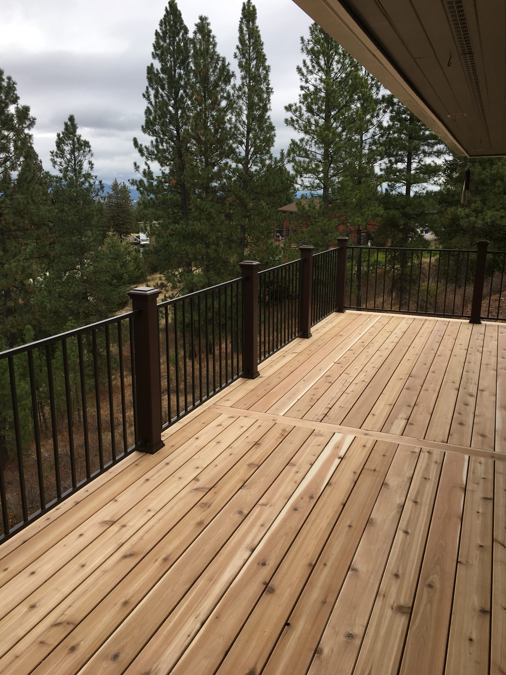 New cedar wood deck with black picket railing under roof overhang, pine trees behind