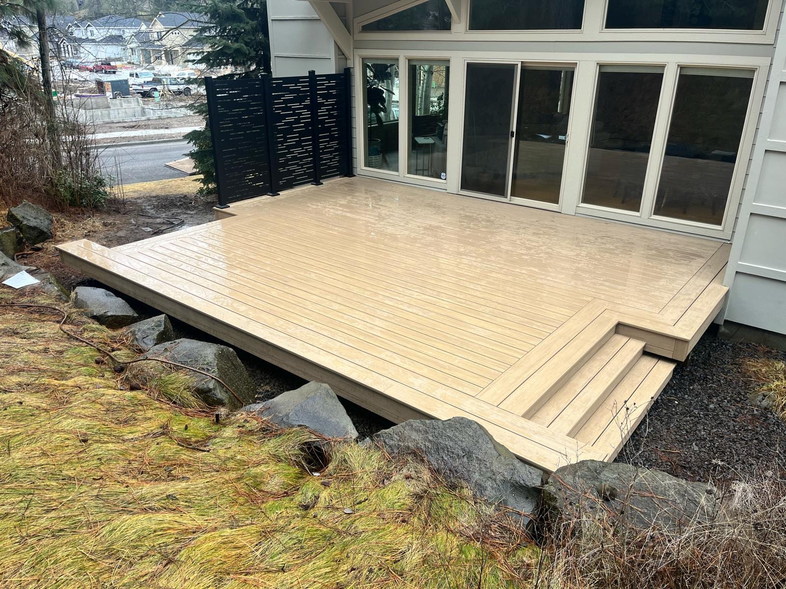 New cream composite low deck with two-step corner beside grey house with large windows, black privacy screen, rocks and grass