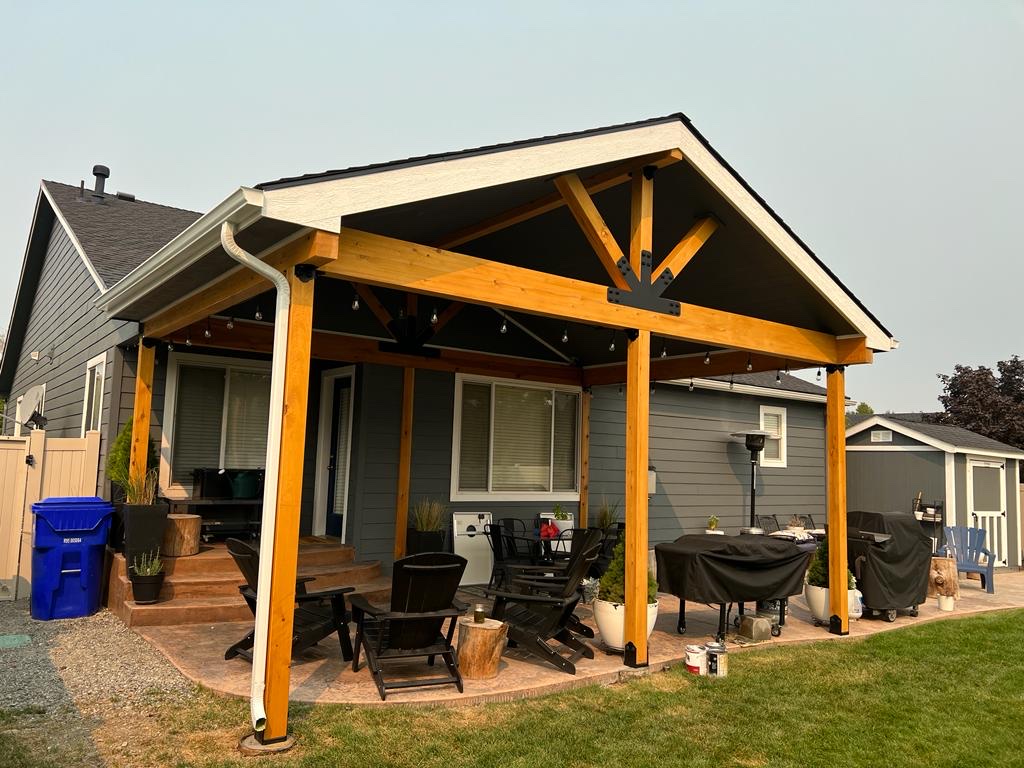New gabled timber-frame cedar patio cover with truss accent over paver patio and outdoor furniture, hazy sky