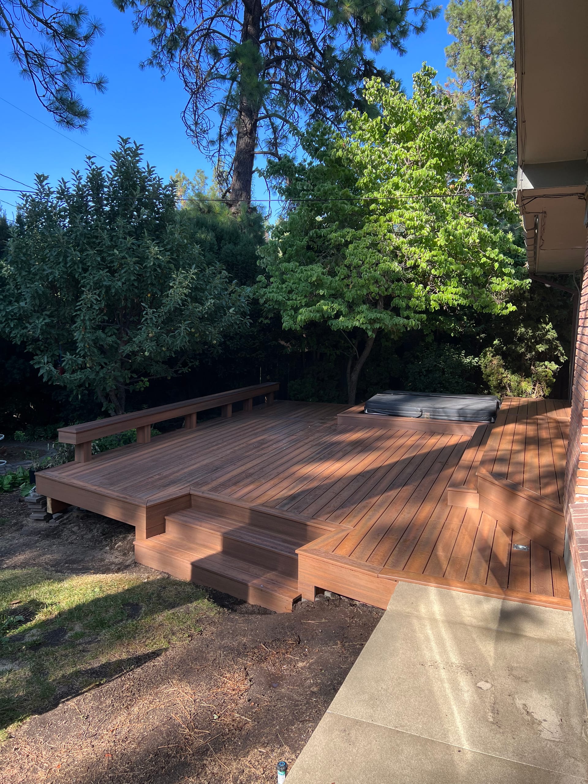 New red-brown hardwood deck with steps and built-in bench among trees, hot tub partly visible