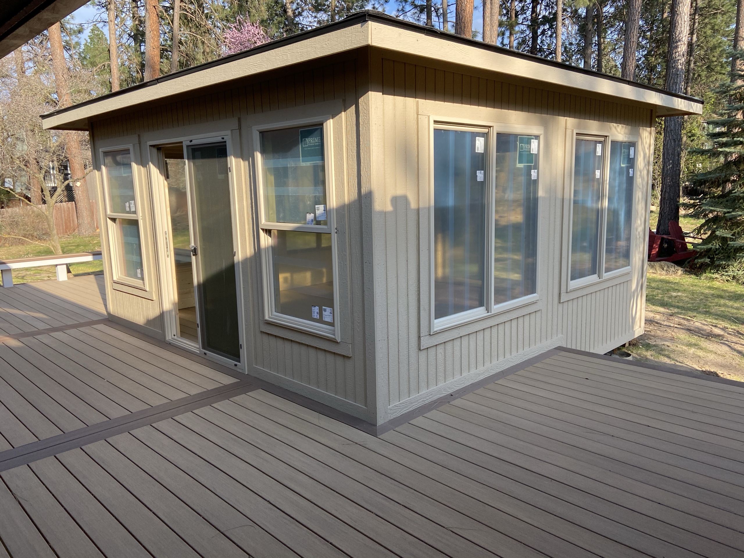 New small enclosed tan-sided room with windows on a large tan composite deck with brown border, pine trees behind