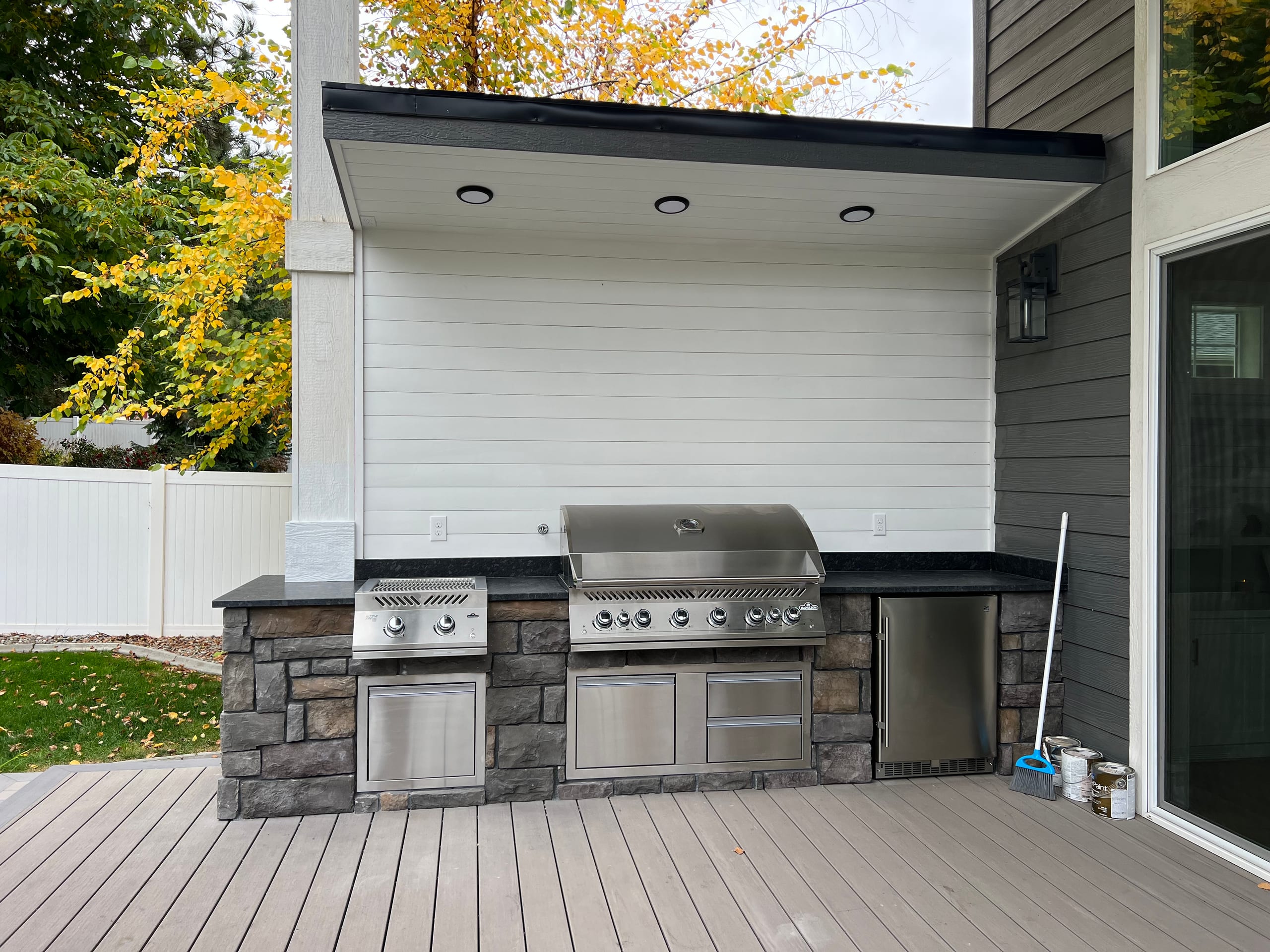 Outdoor kitchen with stone base, stainless grill and fridge under a covered white alcove on gray composite deck, autumn tree