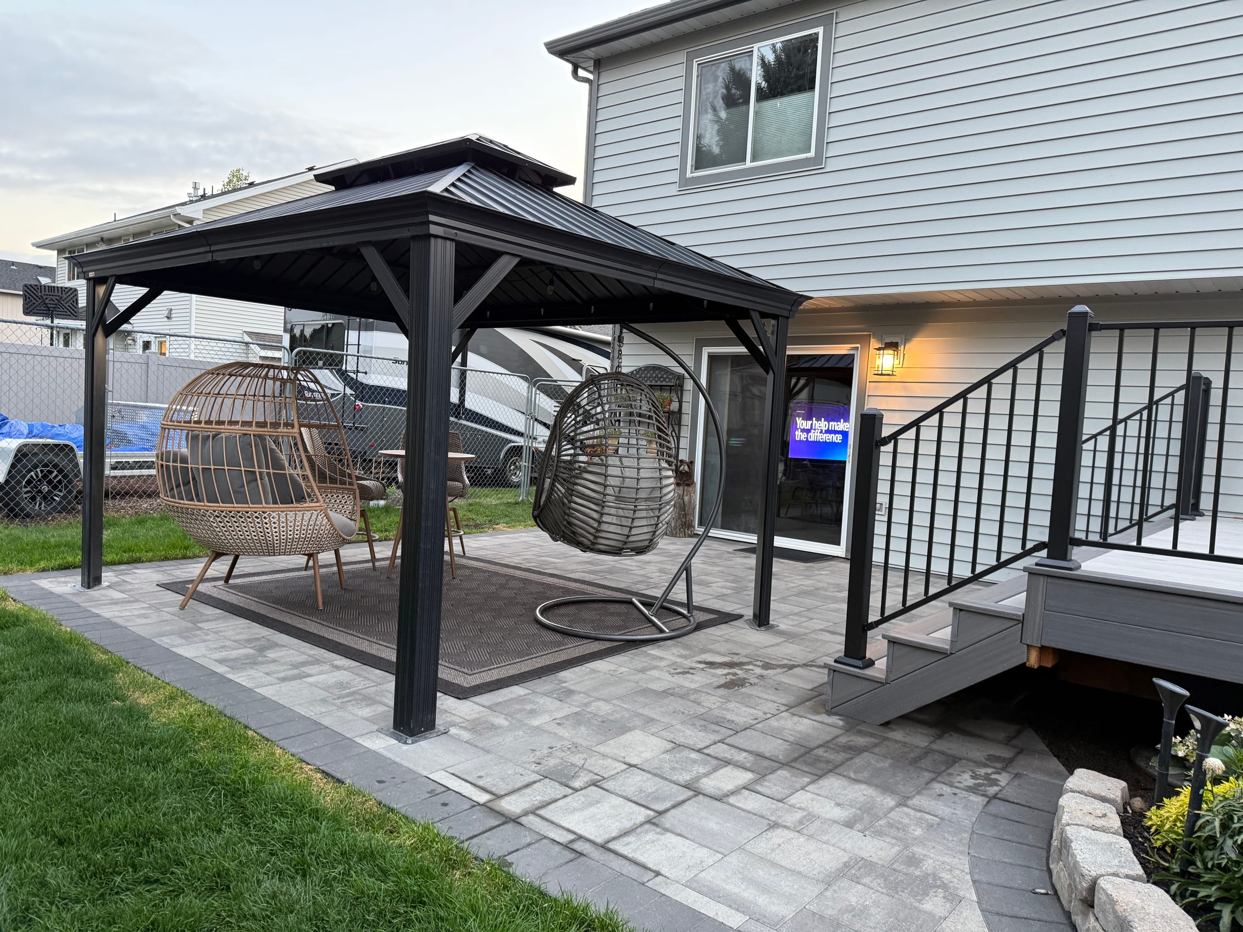 Paver patio with black gazebo, wicker egg chairs, deck steps and black railing, flower bed at edge, evening light