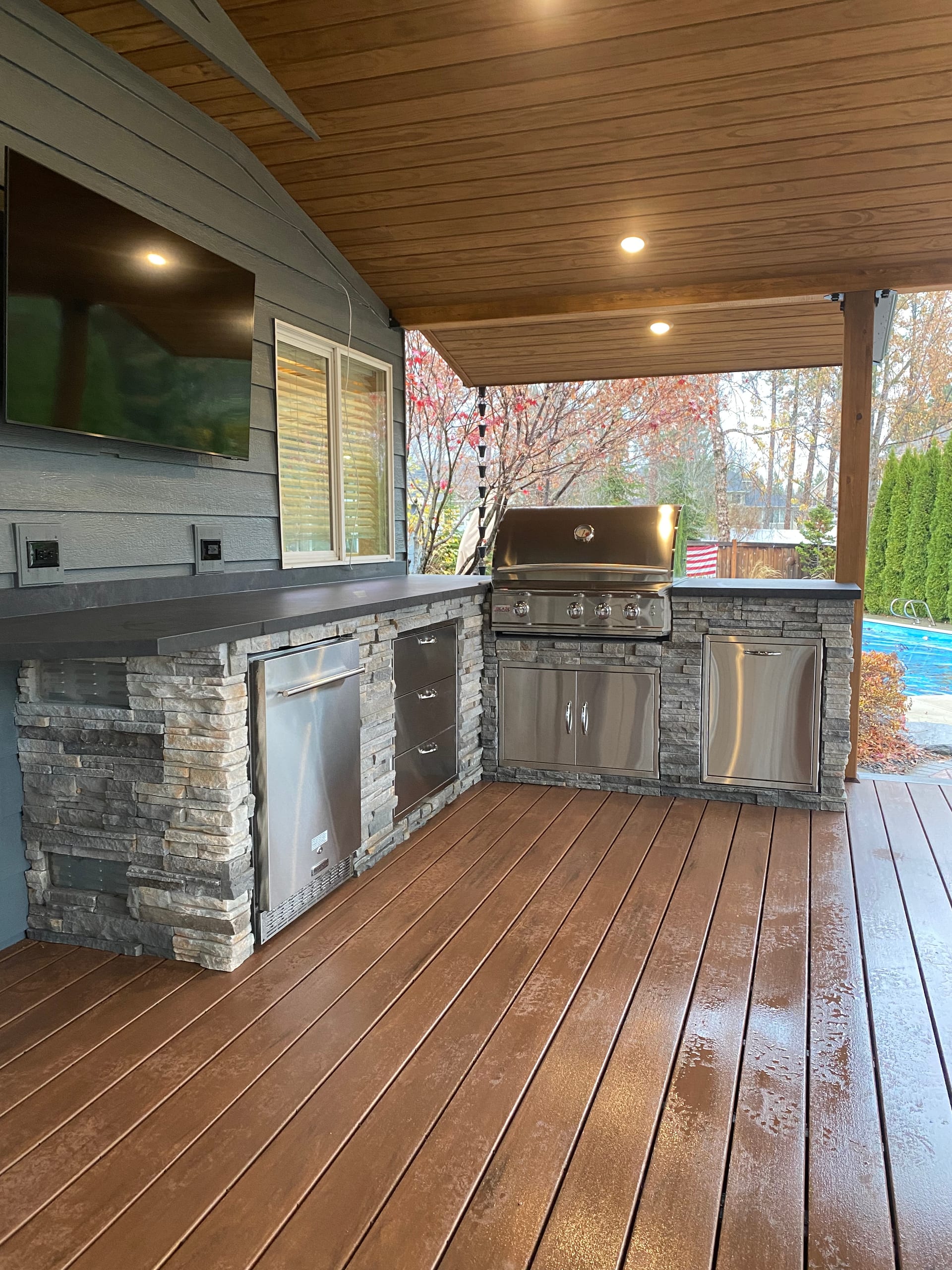 Portrait of outdoor kitchen run along gray wall with TV and stainless grill on a wet brown composite deck