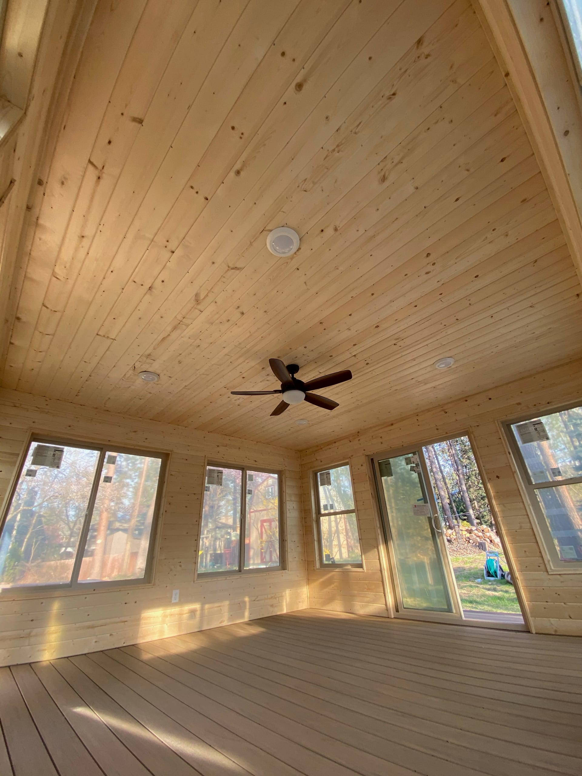 Portrait view of a vaulted wood-ceiling enclosed room with ceiling fan and windows on all sides, light plank floor