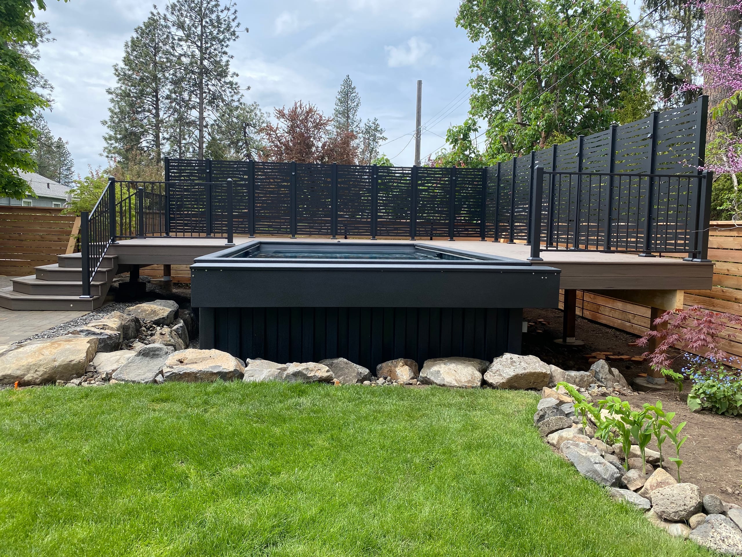 Raised deck with black slatted privacy screens above a rectangular pool and rock border