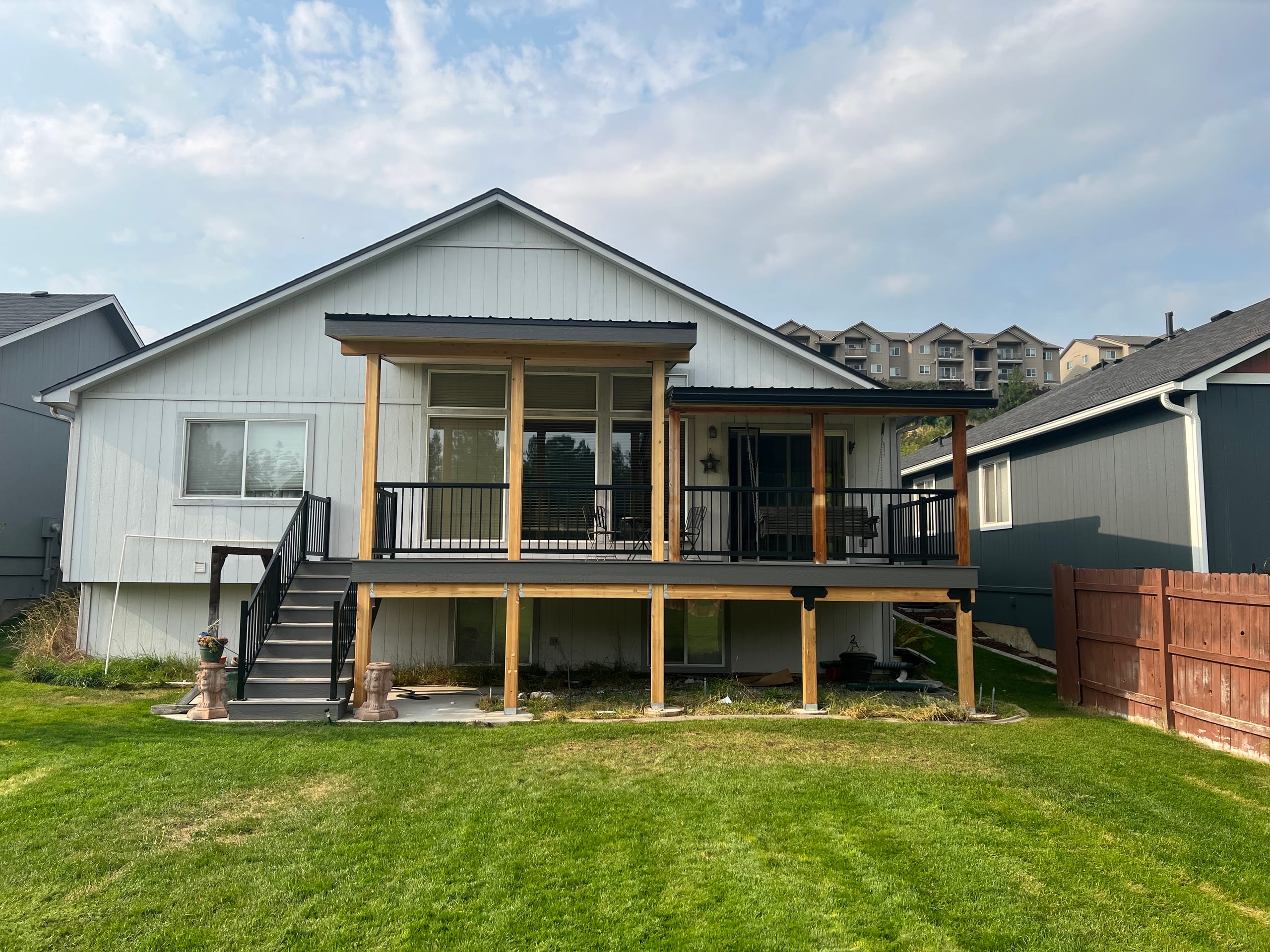 Raised deck with stained-timber glassy upper room or cover, black railing and stairs on a gray house, mowed lawn