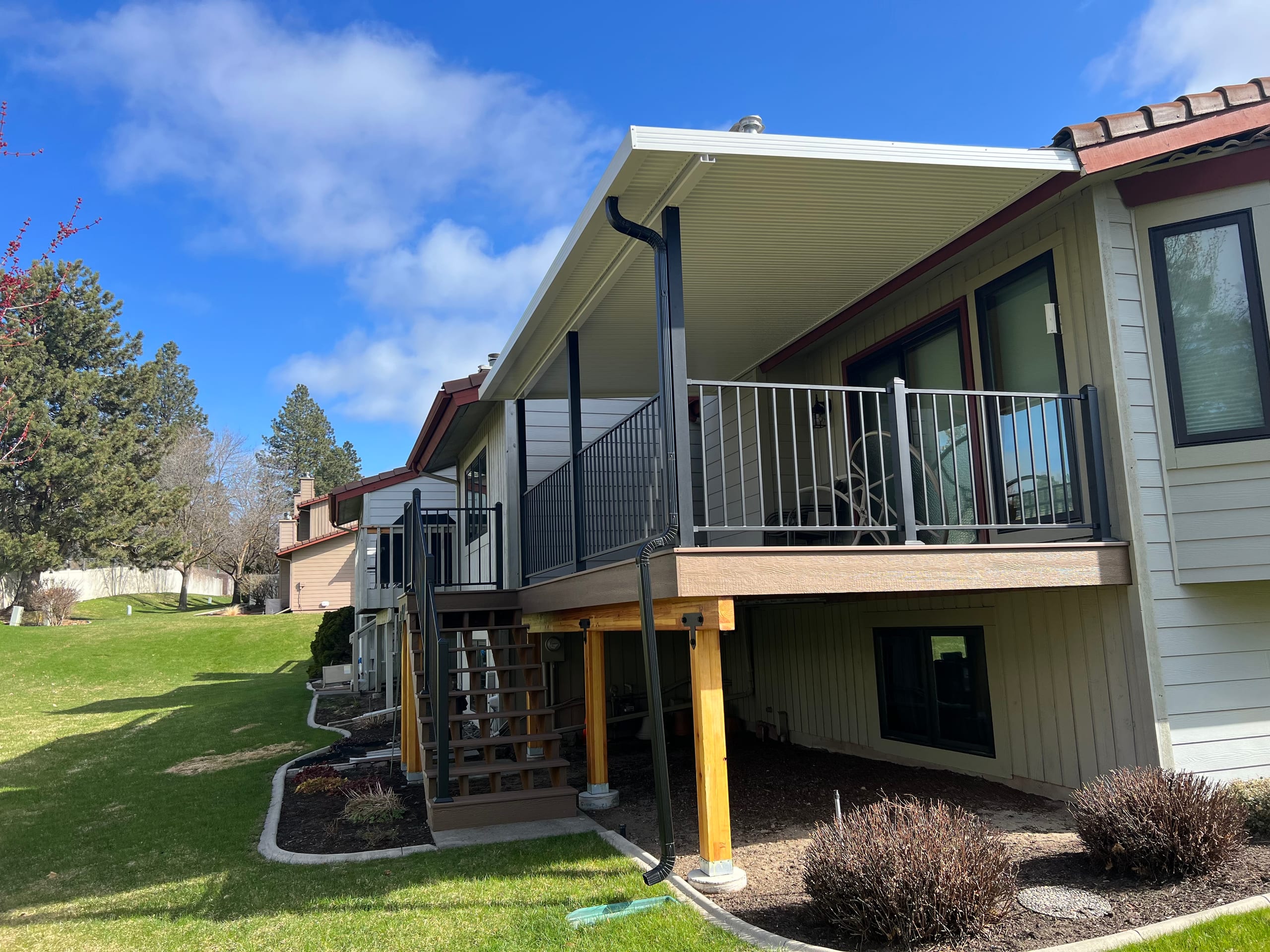 Raised deck with white metal-panel roof cover, black railing and stained posts below on beige two-story house, green lawn
